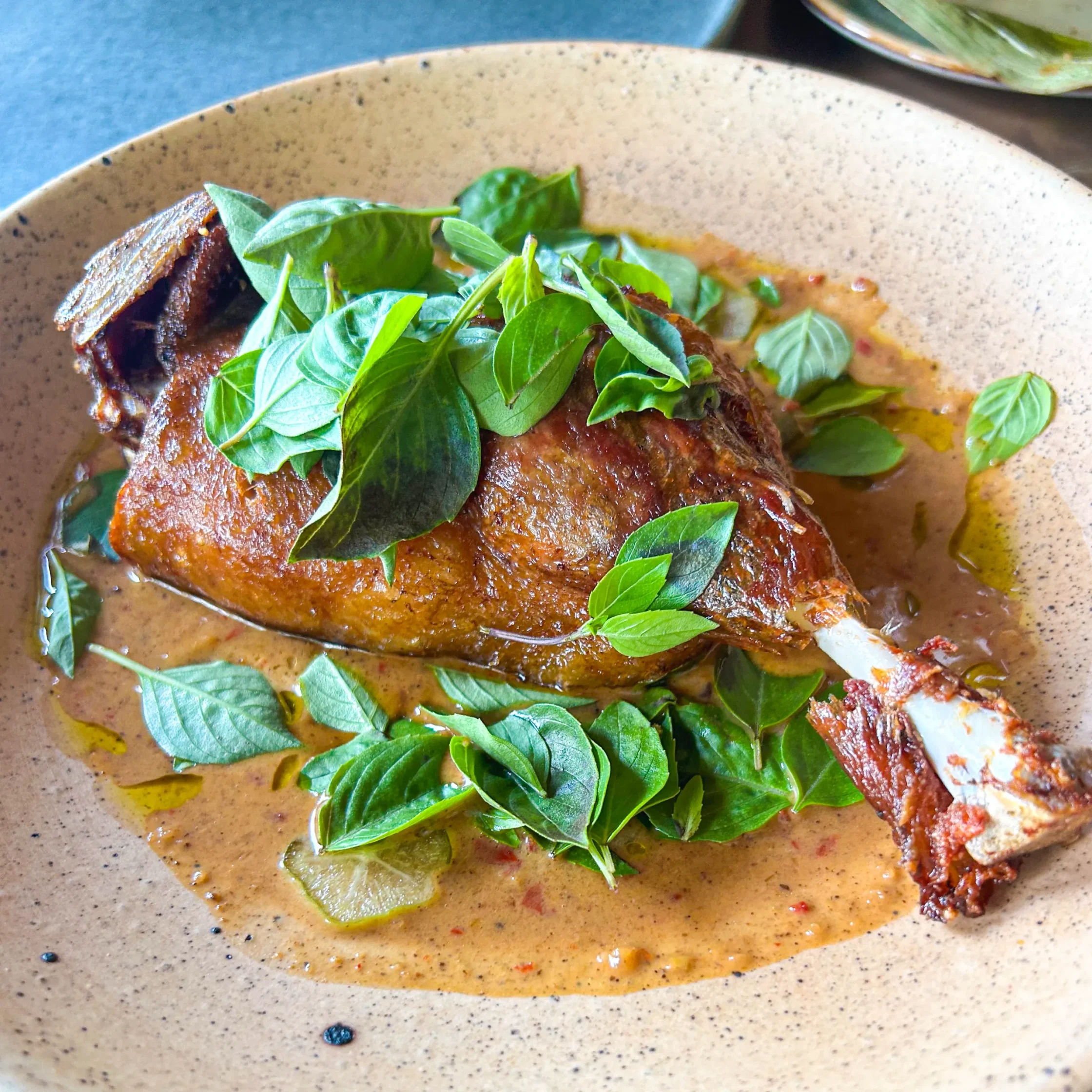Golden crispy duck leg resting in a creamy pineapple red curry sauce, generously garnished with fresh herbs and served in a speckled ceramic bowl.