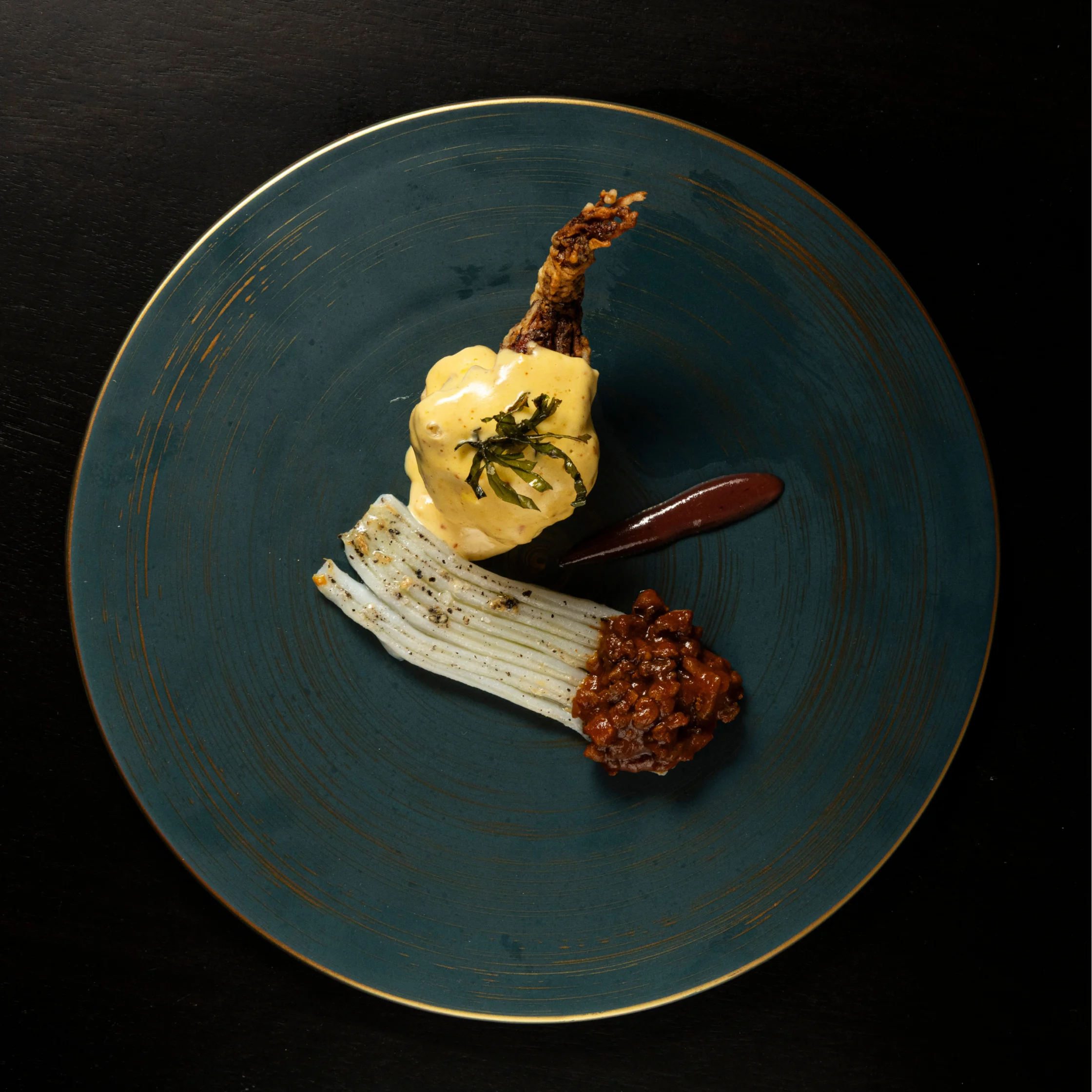 Plated fine-dining dish on a dark blue ceramic plate featuring a crispy vegetable centerpiece topped with creamy yellow sauce, grilled white vegetable stalks, a dark sauce smear, and a savory relish garnish.
