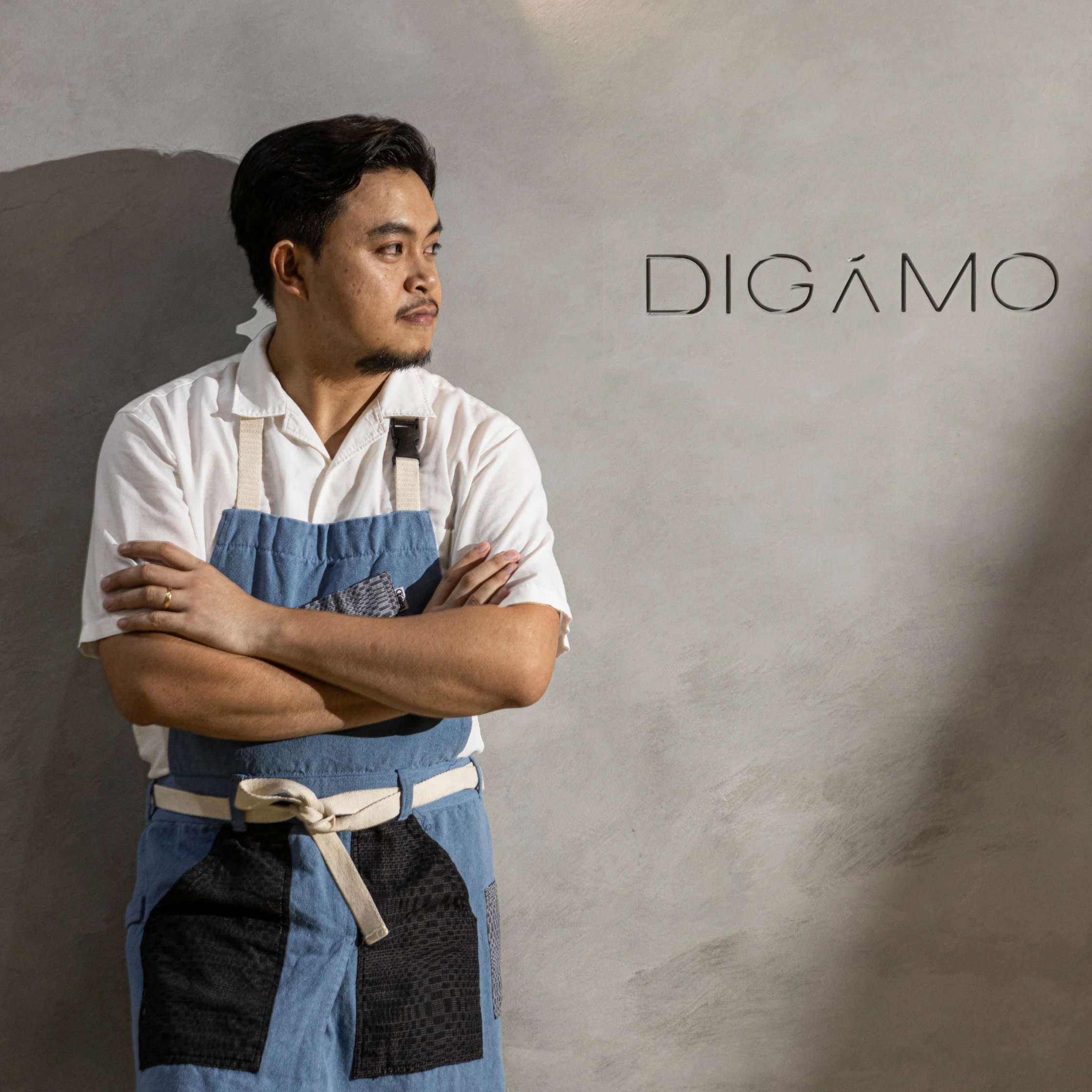 Chef Francis Lacson standing with arms crossed in a blue apron against a minimalist wall with the DIGAMO restaurant logo, looking confidently to the side.