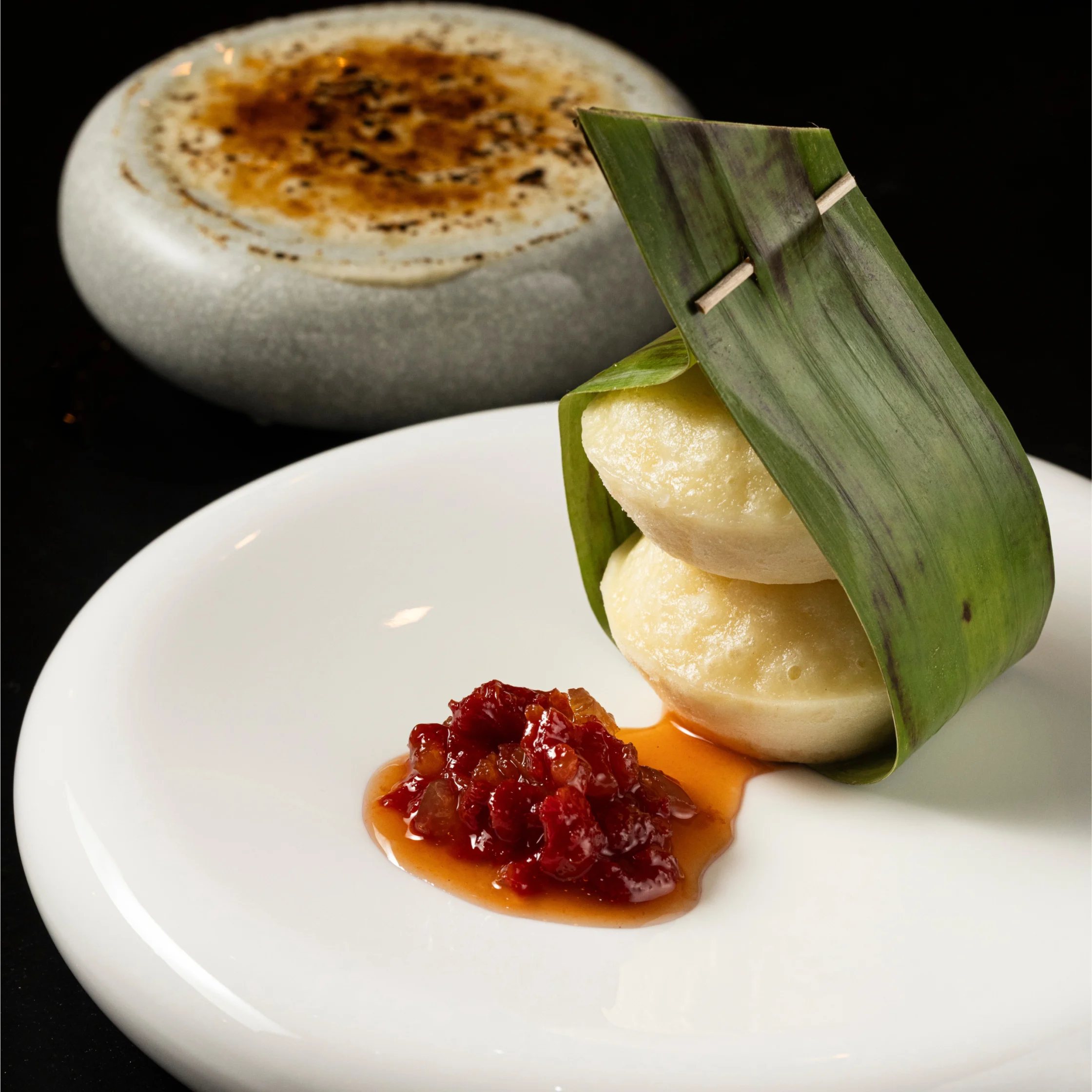 Two steamed rice cakes wrapped in a folded banana leaf and secured with a wooden pin, served with a glossy red fruit preserve on a white plate.