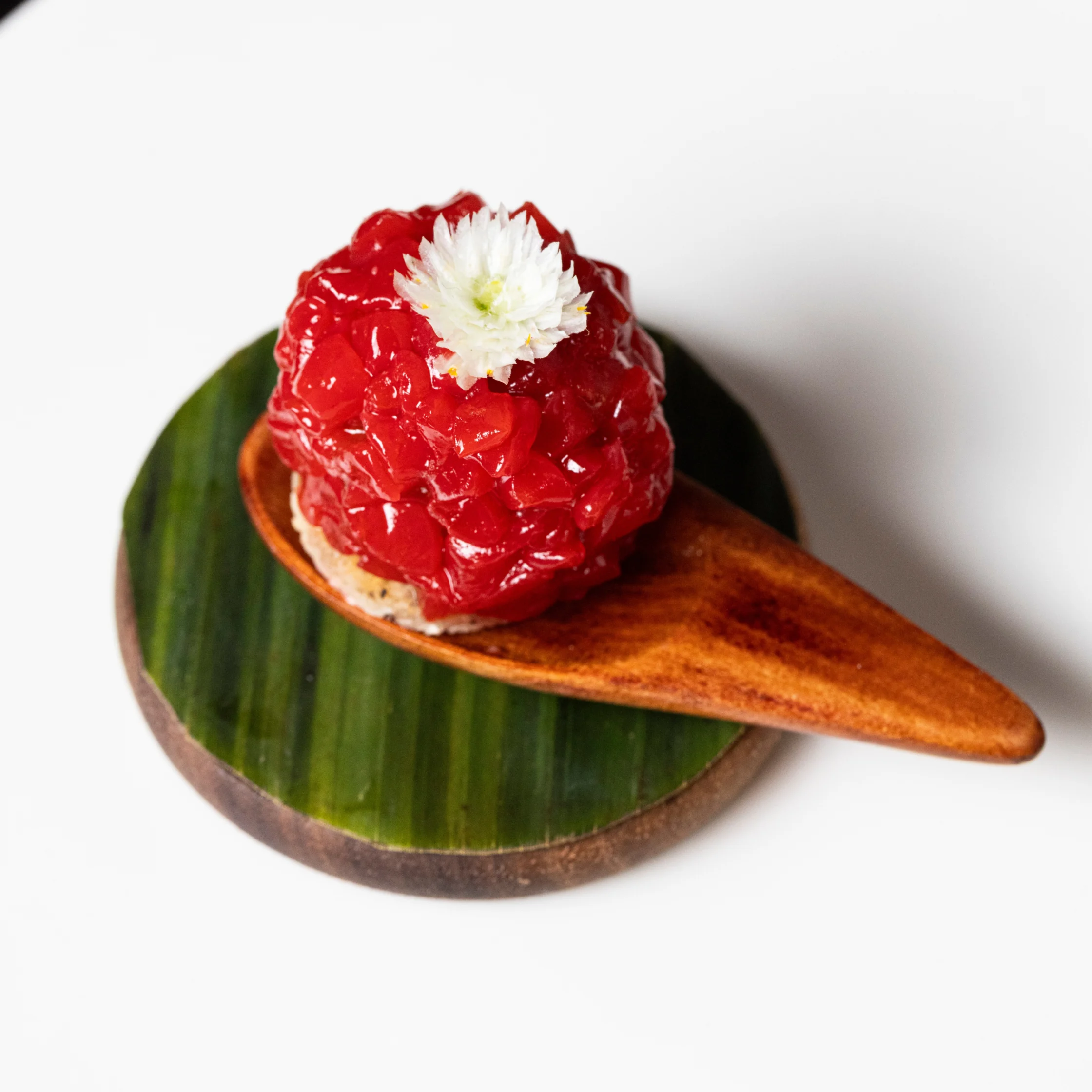 Bright red nilupak-style dessert topped with a small white edible flower, served on a wooden spoon resting on a round banana-leaf base.