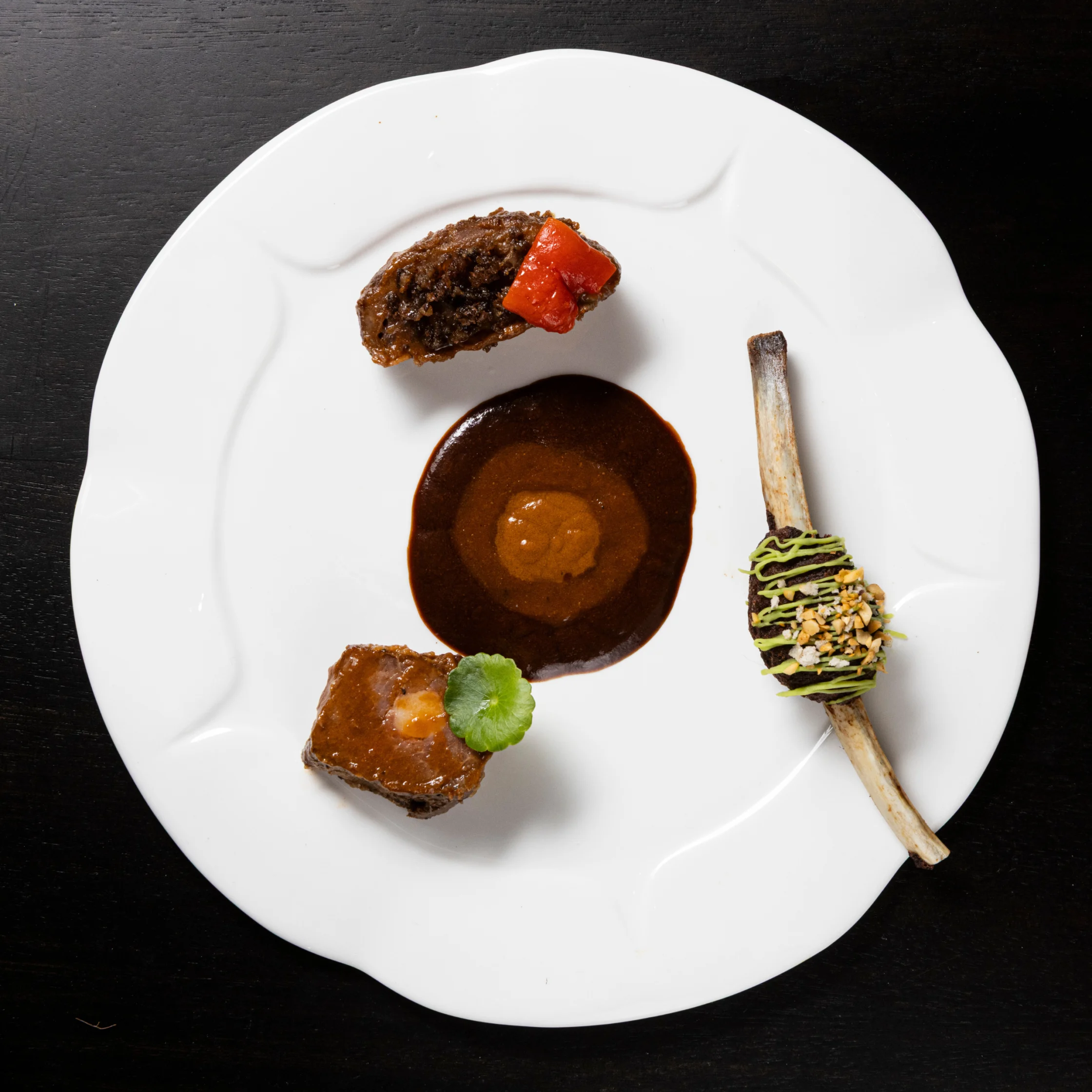 Contemporary plated meat course featuring three components arranged around a rich brown sauce, including a bone-in preparation garnished with herbs and crushed nuts.