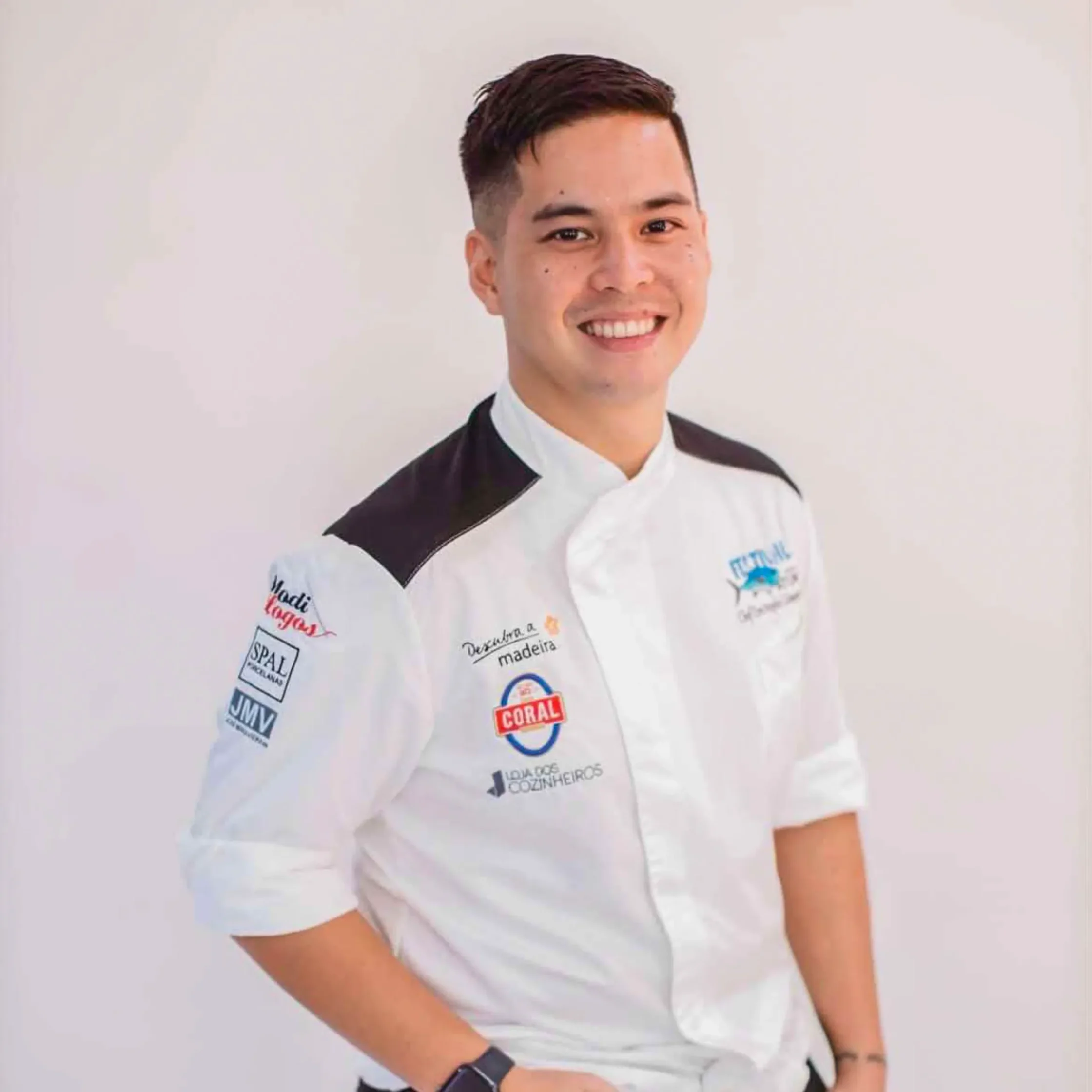 Portrait of chef don colmenares in a white culinary jacket with sponsor logos, standing against a plain light background.