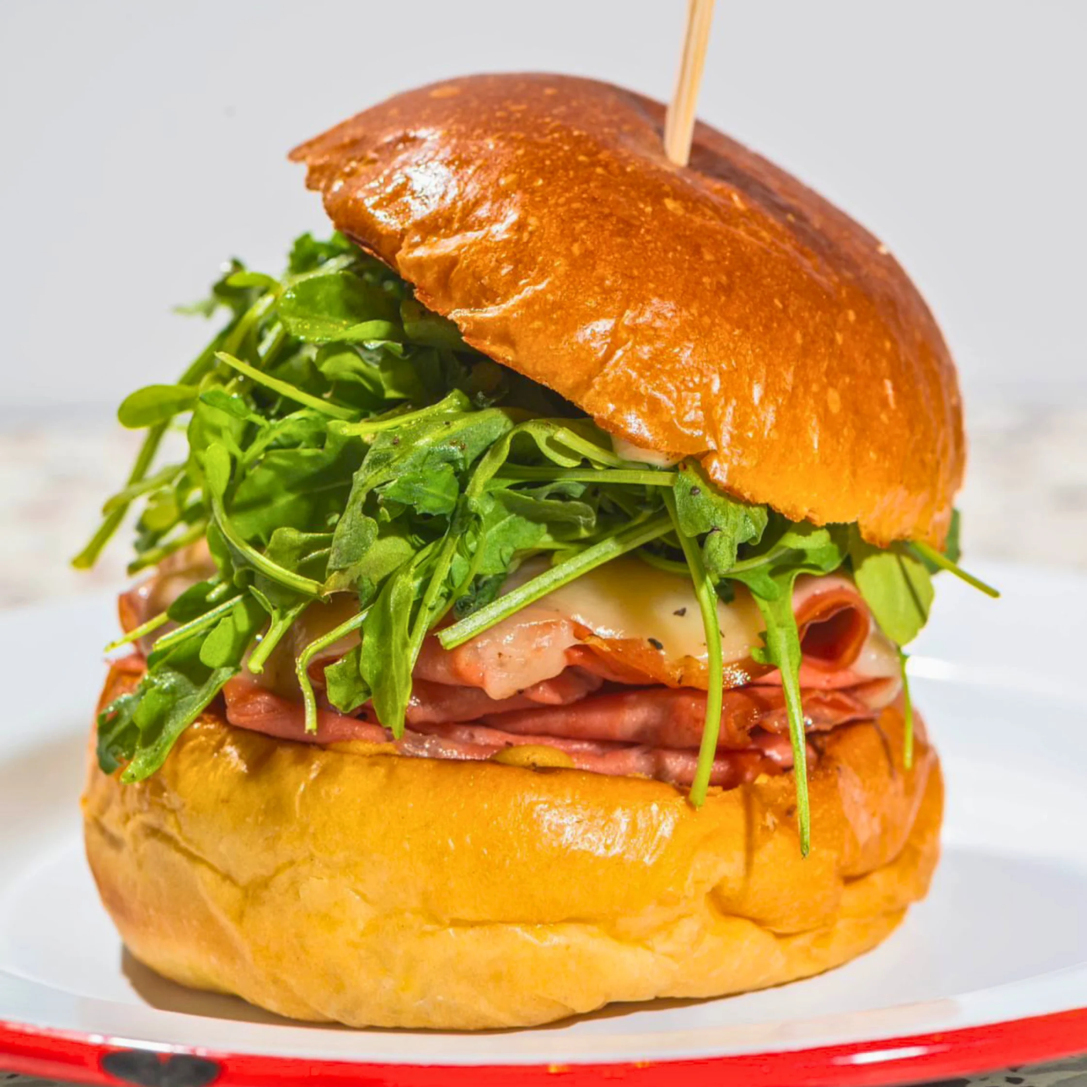 Close-up of a brioche bun sandwich filled with seared mortadella, melted provolone cheese, and a generous pile of fresh arugula, secured with a skewer and served on a plate.