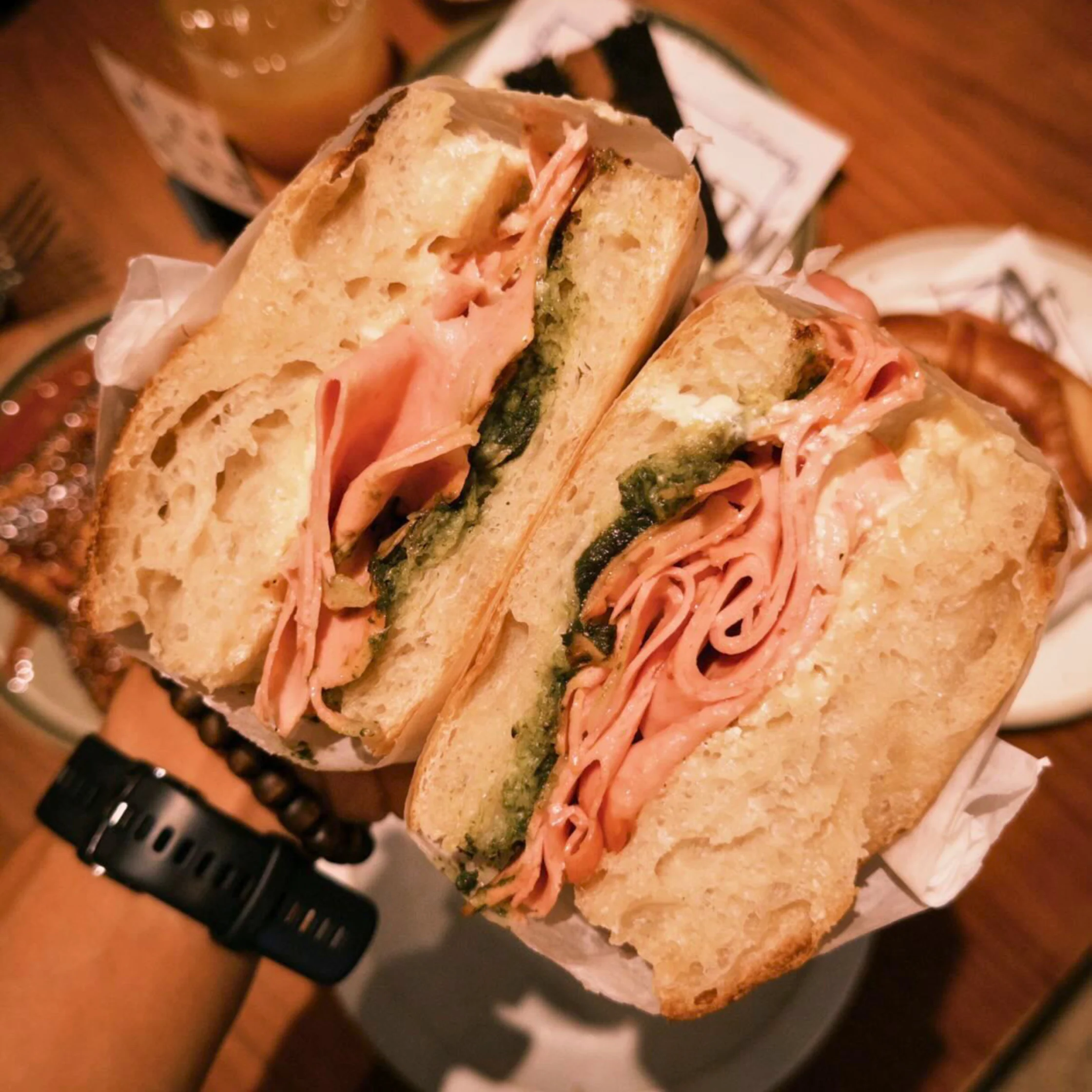 Handheld bourdain sandwich cut in half, revealing layers of sliced mortadella, pesto, and whipped ricotta, photographed over a restaurant table.