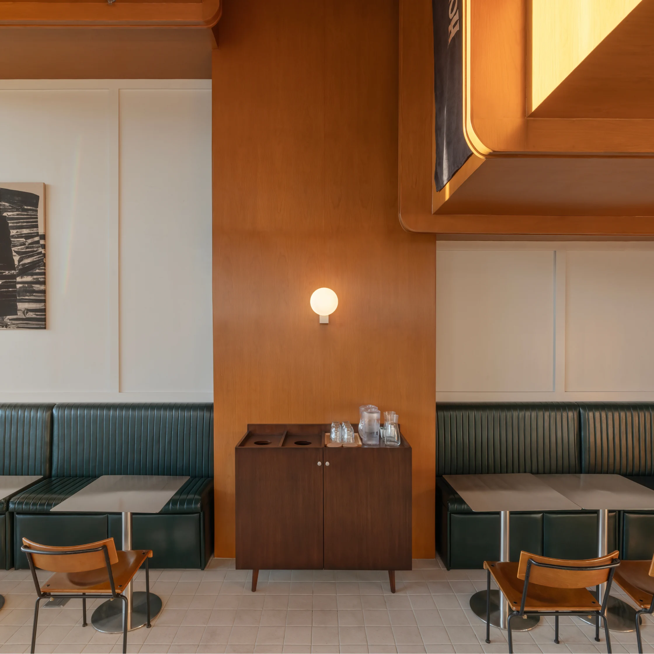 Interior of Hometown Cafe featuring green booth seating, warm wood paneling, small dining tables, and a mid-century style cabinet beneath a glowing wall light.