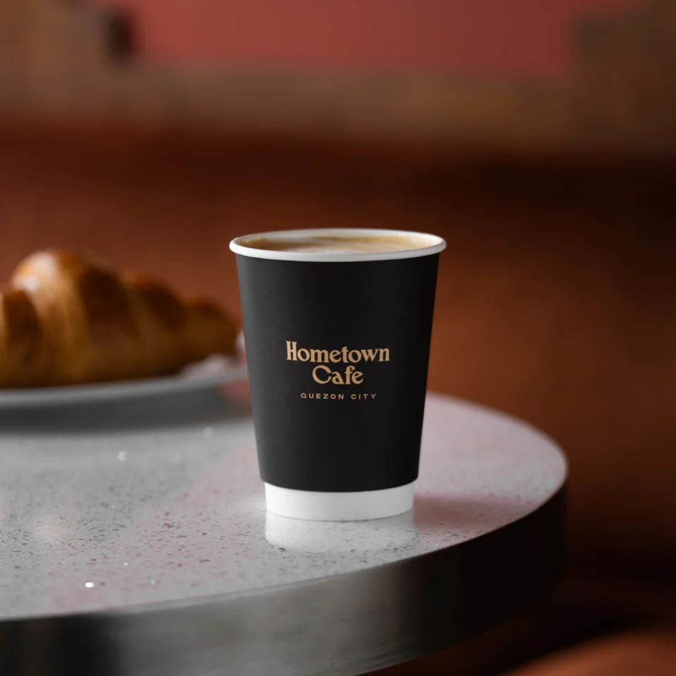 Branded Hometown Cafe takeaway coffee cup on a table, with a croissant softly blurred in the background.