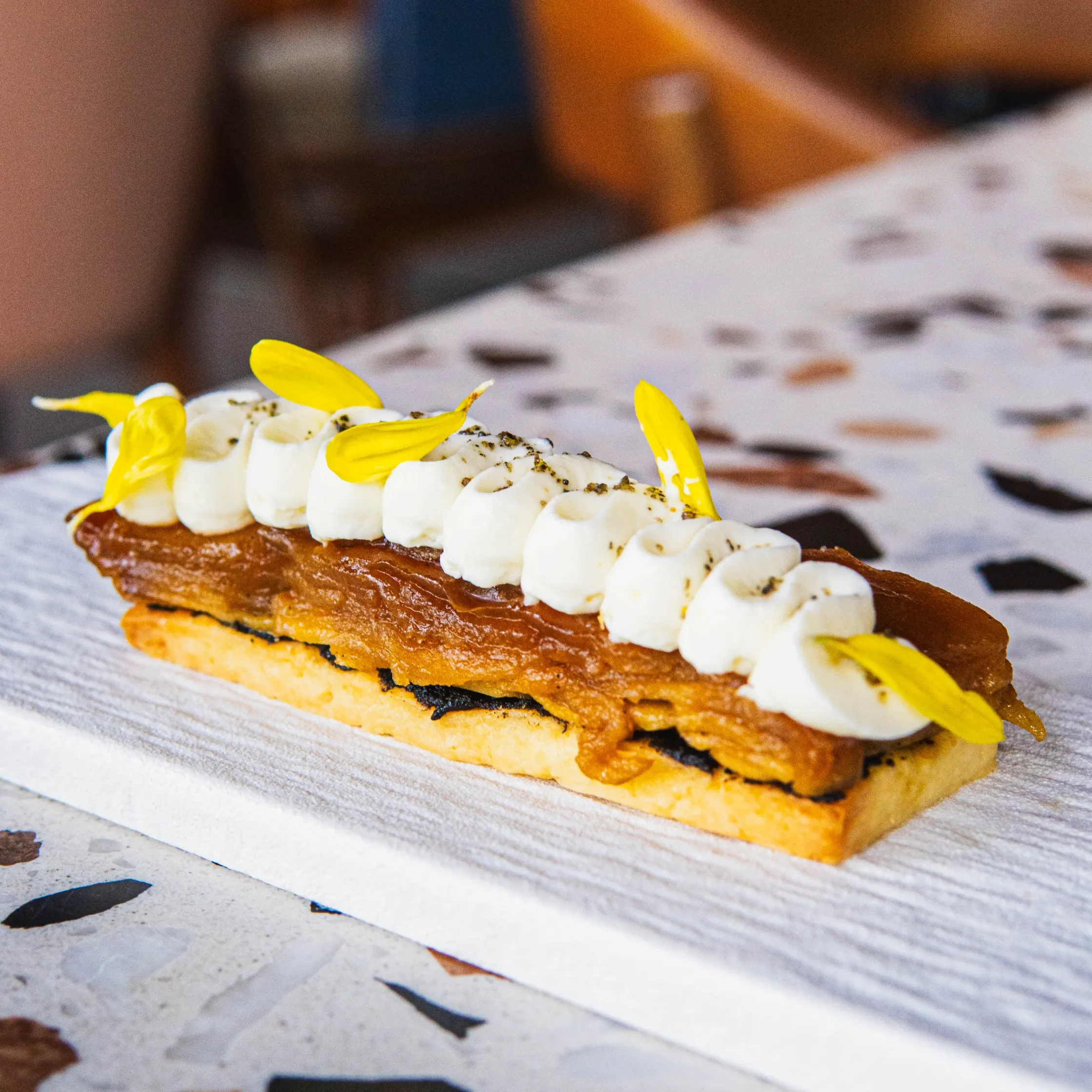 Kurogoma tarte tatin topped with piped cream and yellow edible flower petals on a rectangular serving plate.