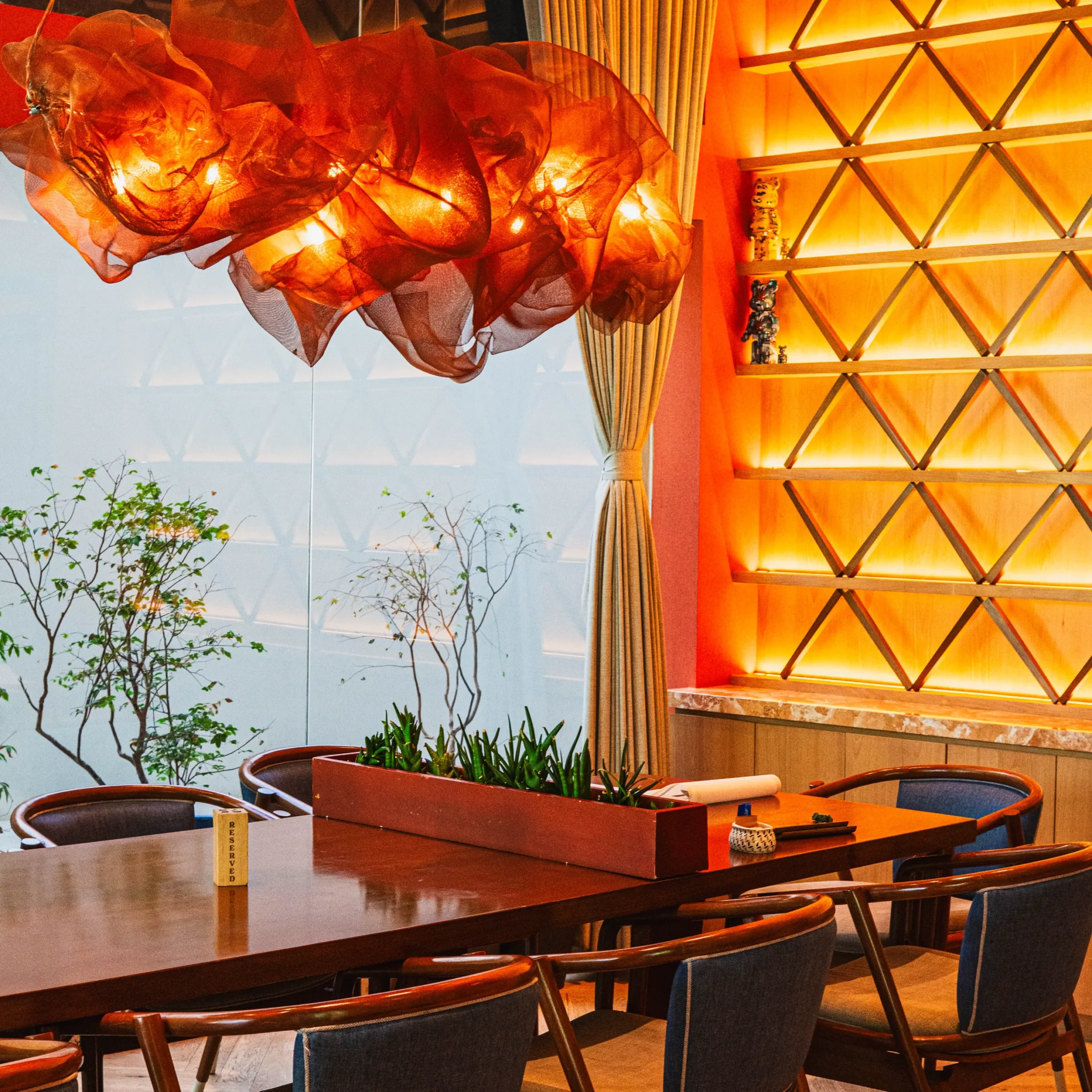 Modern KAZU Café dining area featuring a long wooden communal table, upholstered chairs, warm amber lighting, a dramatic red sculptural fabric chandelier, geometric backlit wall shelving, and floor-to-ceiling windows overlooking a landscaped garden.
