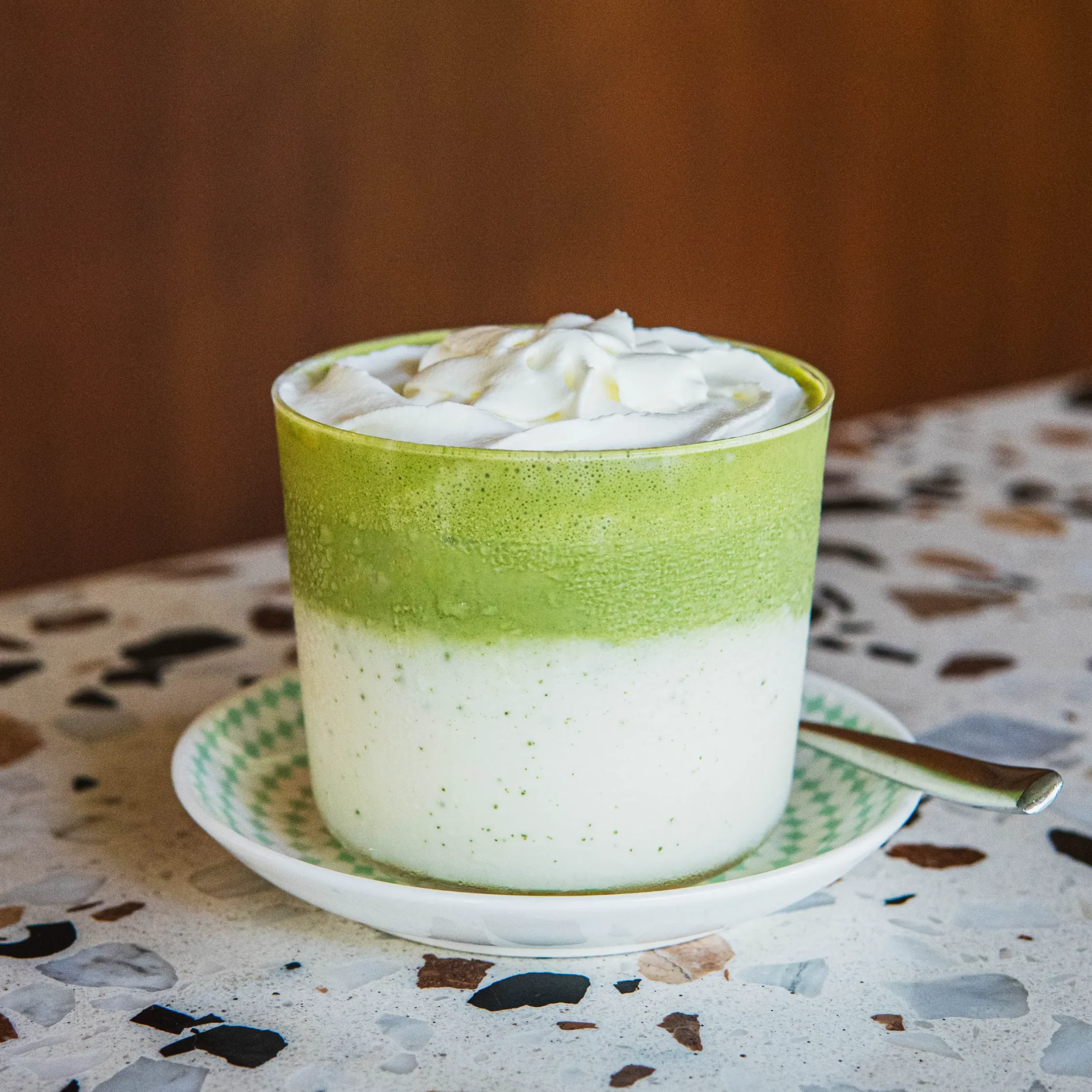 Layered iced matcha and milk drink topped with whipped cream, served in a glass on a patterned saucer.