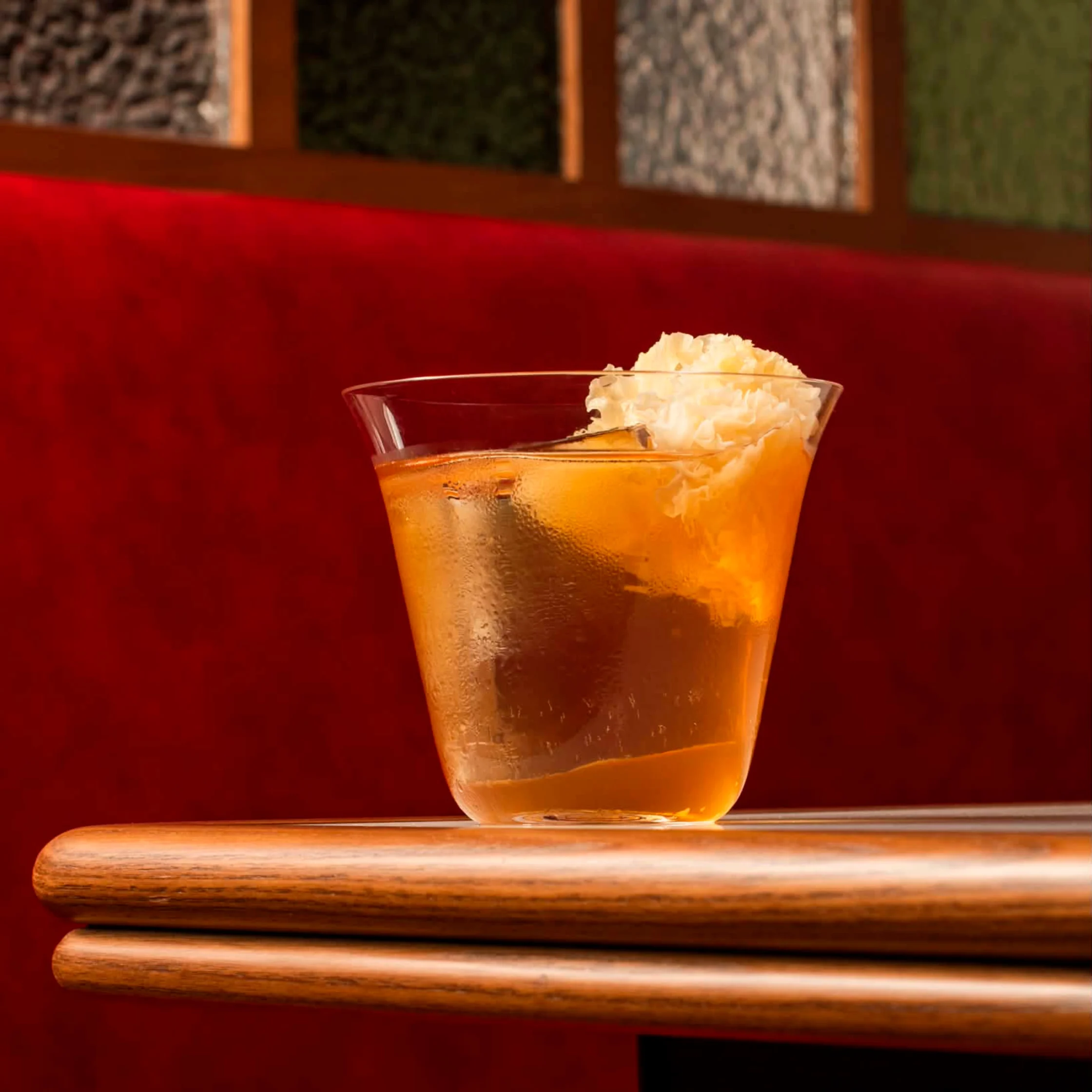 A golden cocktail topped with a delicate garnish rests on a table against a vibrant red banquette, highlighting one of the specially created drinks for Kinsman and Lazy Suzy’s two-night collaboration.