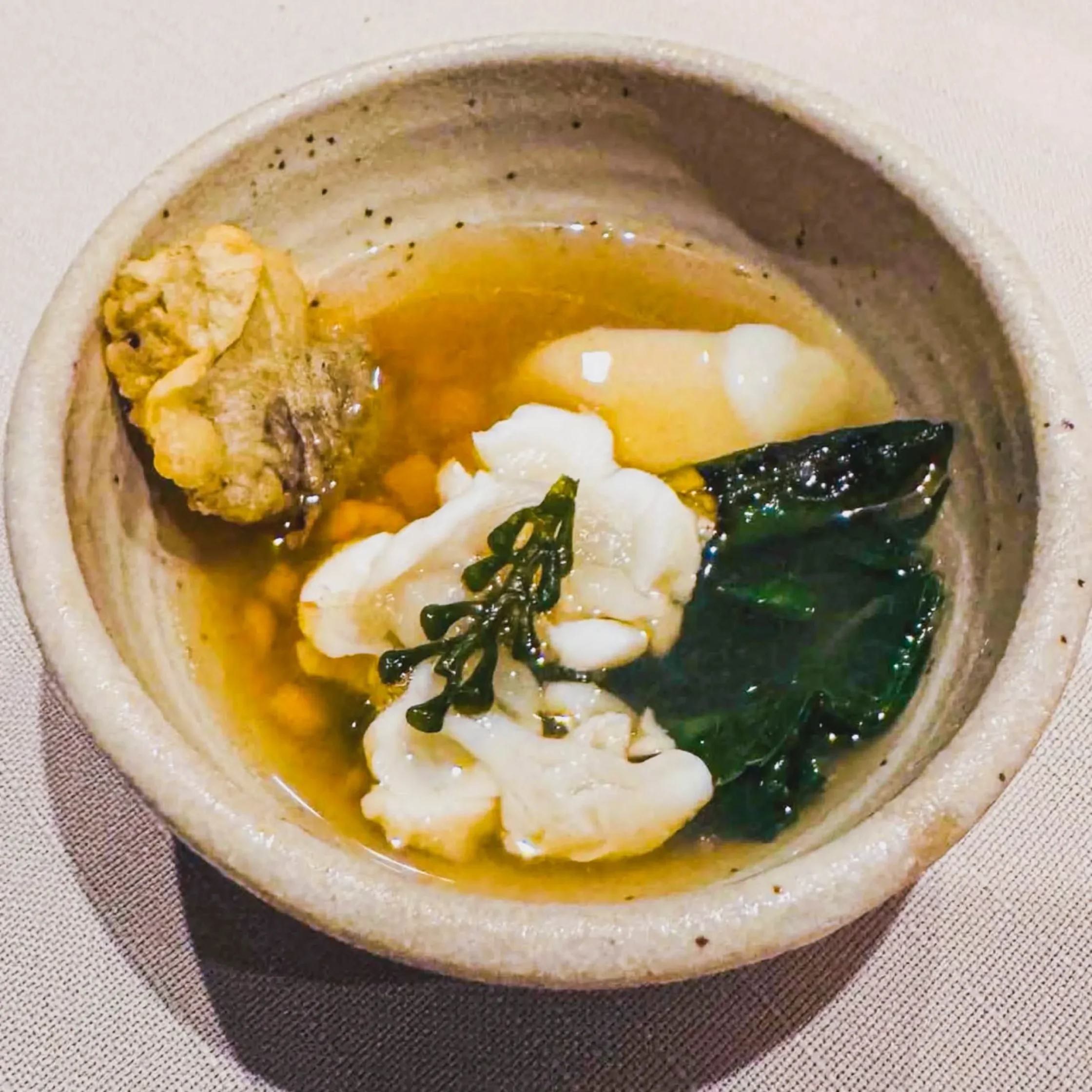 Small ceramic bowl of lobster laswa soup with lobster meat, leafy greens, and vegetables in a golden broth.