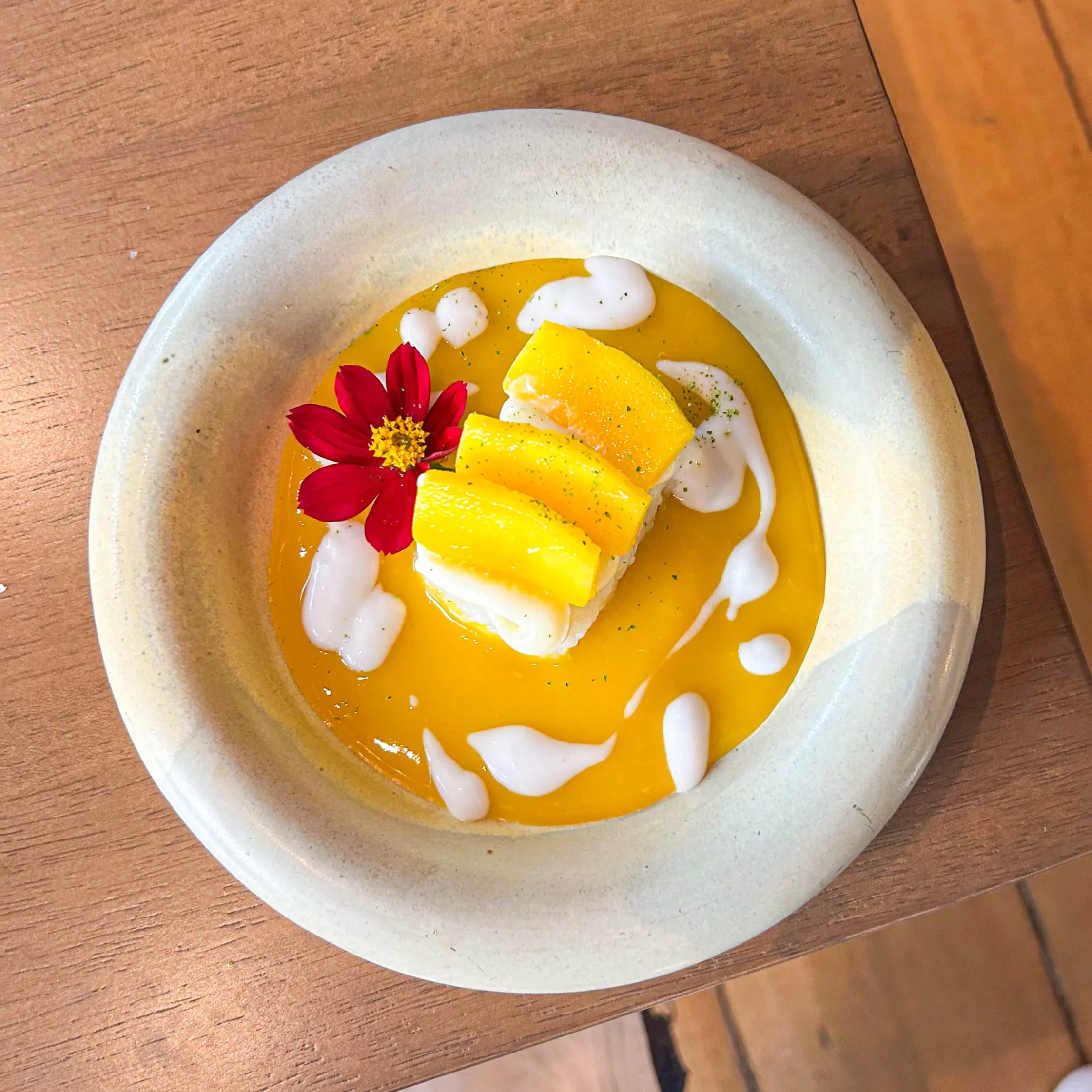 A plated mango dessert featuring slices of ripe mango over coconut rice, surrounded by mango sauce and coconut cream, garnished with a vibrant red edible flower.