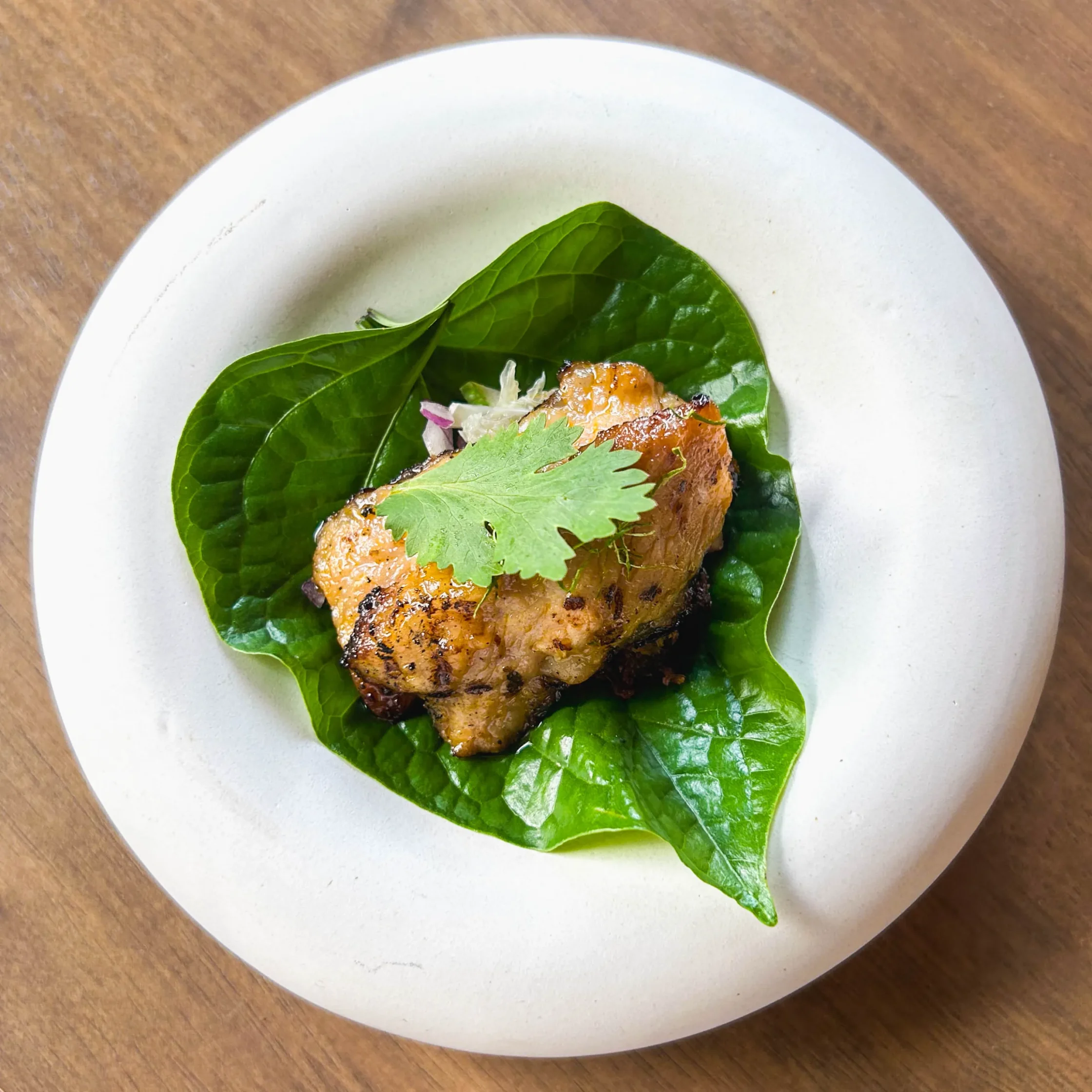 Grilled Iberico pork secreto served atop glossy green leaves with fresh herbs and finely shredded garnish, presented on a minimalist white plate.