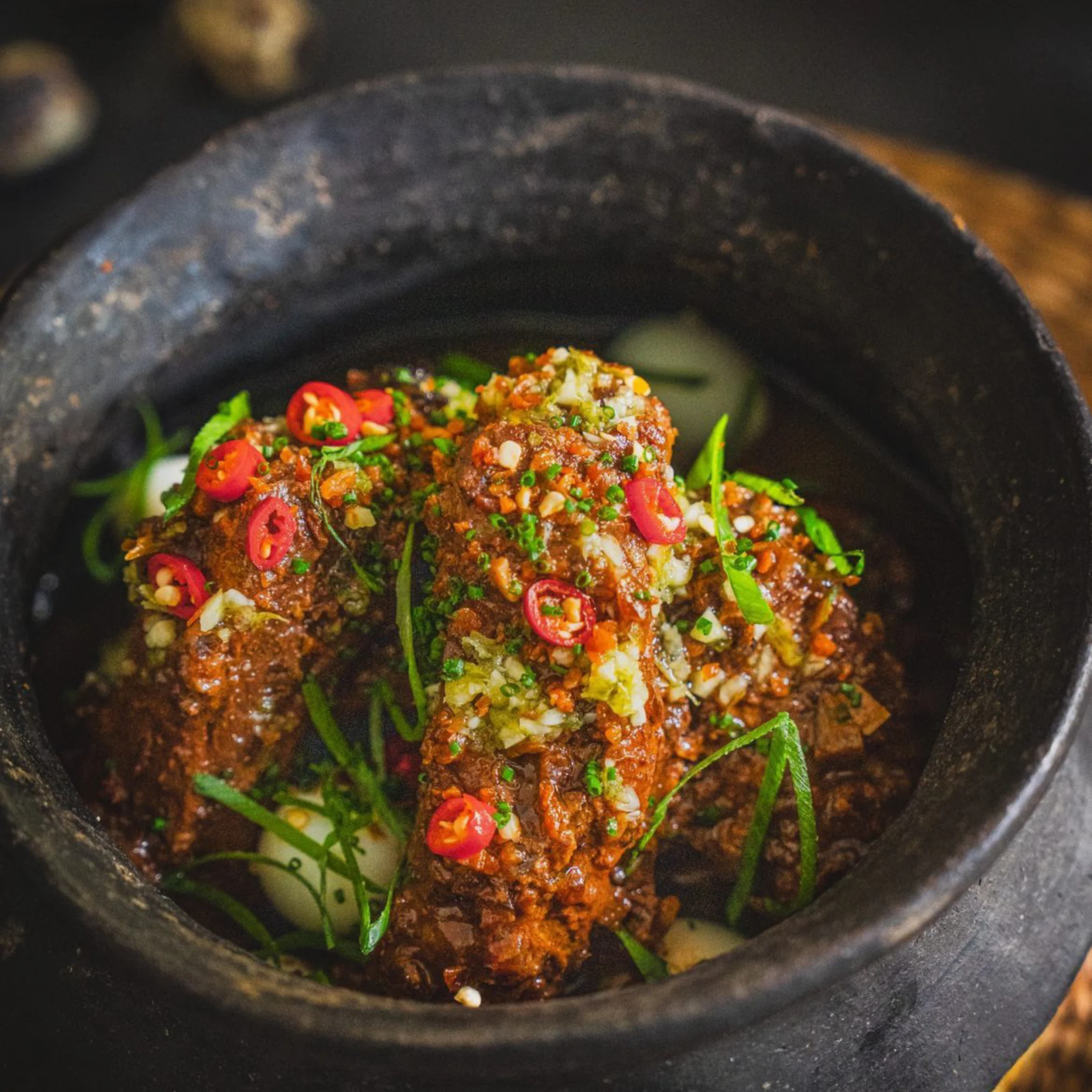 Laguna-style pork rib adobo served in a rustic black clay pot, garnished with sliced red chilies, garlic, and green herbs, with a rich dark sauce coating the meat.