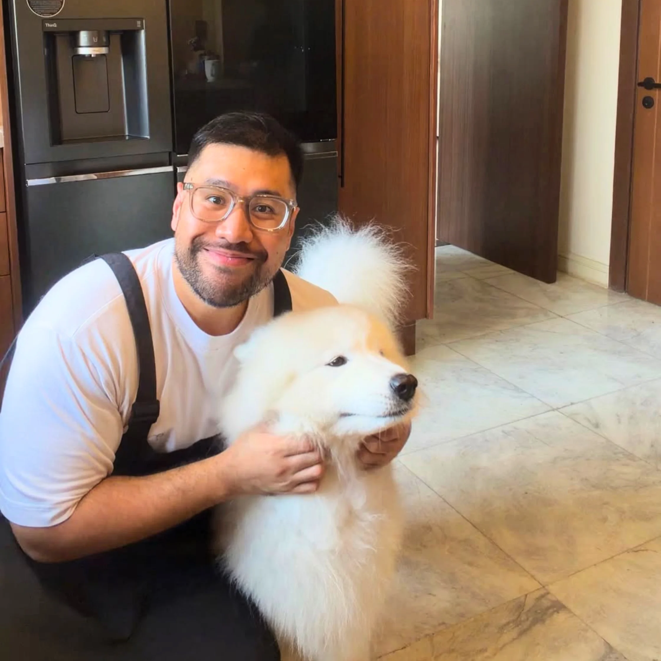 Private Chef Geron Altamirano smiles beside a fluffy white dog indoors, gently holding the dog while posing in a modern home kitchen.