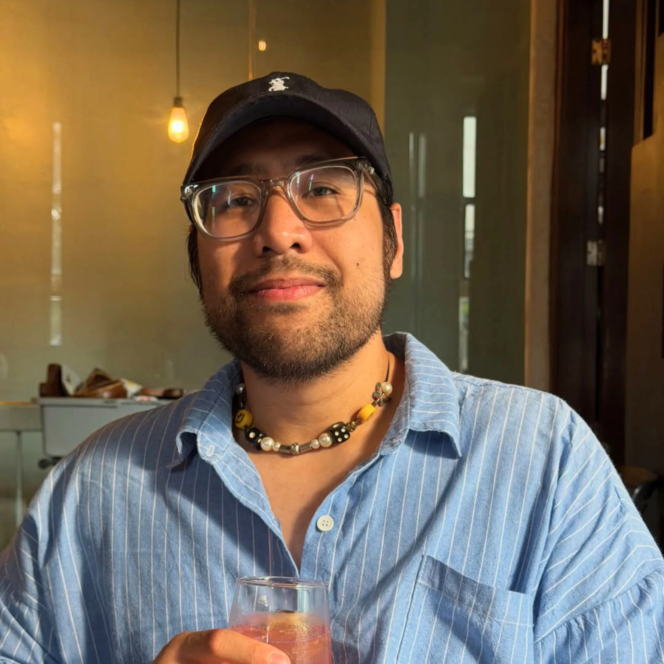 Private Chef Geron Altamirano sits at a table holding a drink, wearing glasses, a black cap, a blue striped shirt, and a beaded necklace under warm indoor lighting.