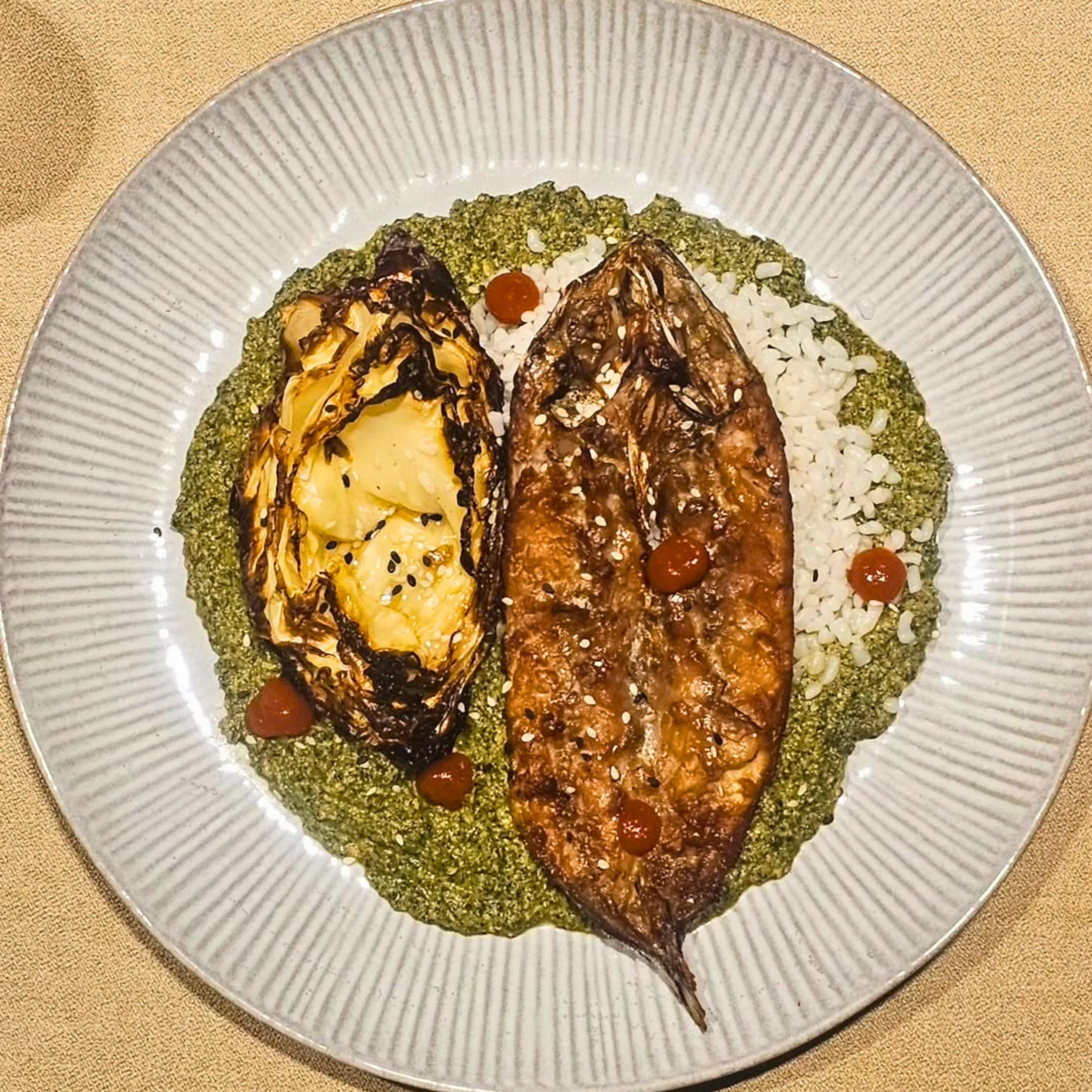 A plated fish entrée by Private Chef Geron Altamirano with grilled fish, roasted cabbage, white rice, green herb sauce, sesame seeds, and red sauce accents.
