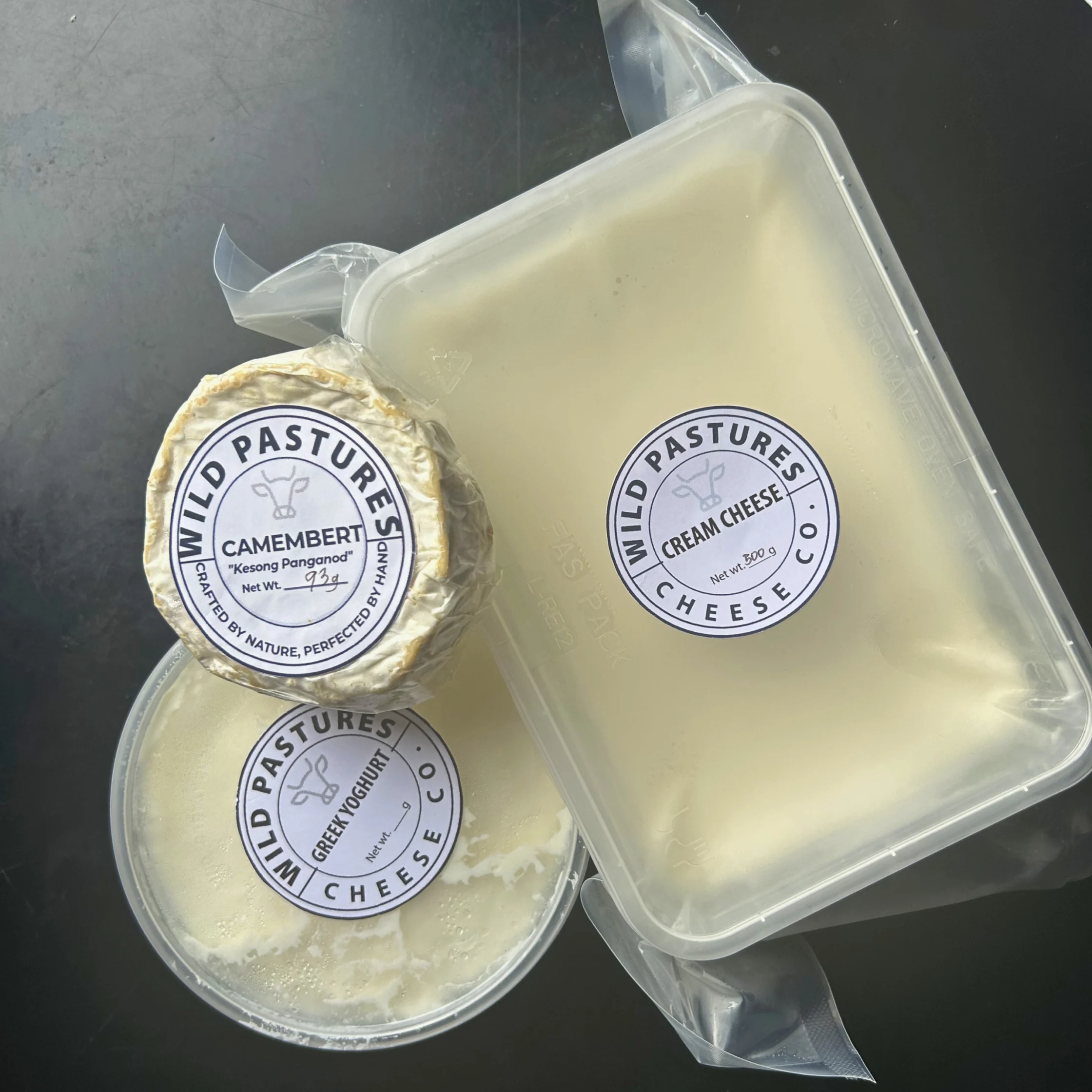 Overhead view of Wild Pastures Cheese products, including Camembert-style cheese, cream cheese, and Greek yogurt in labeled containers from Wild Pasture Cheesemakers on a dark surface