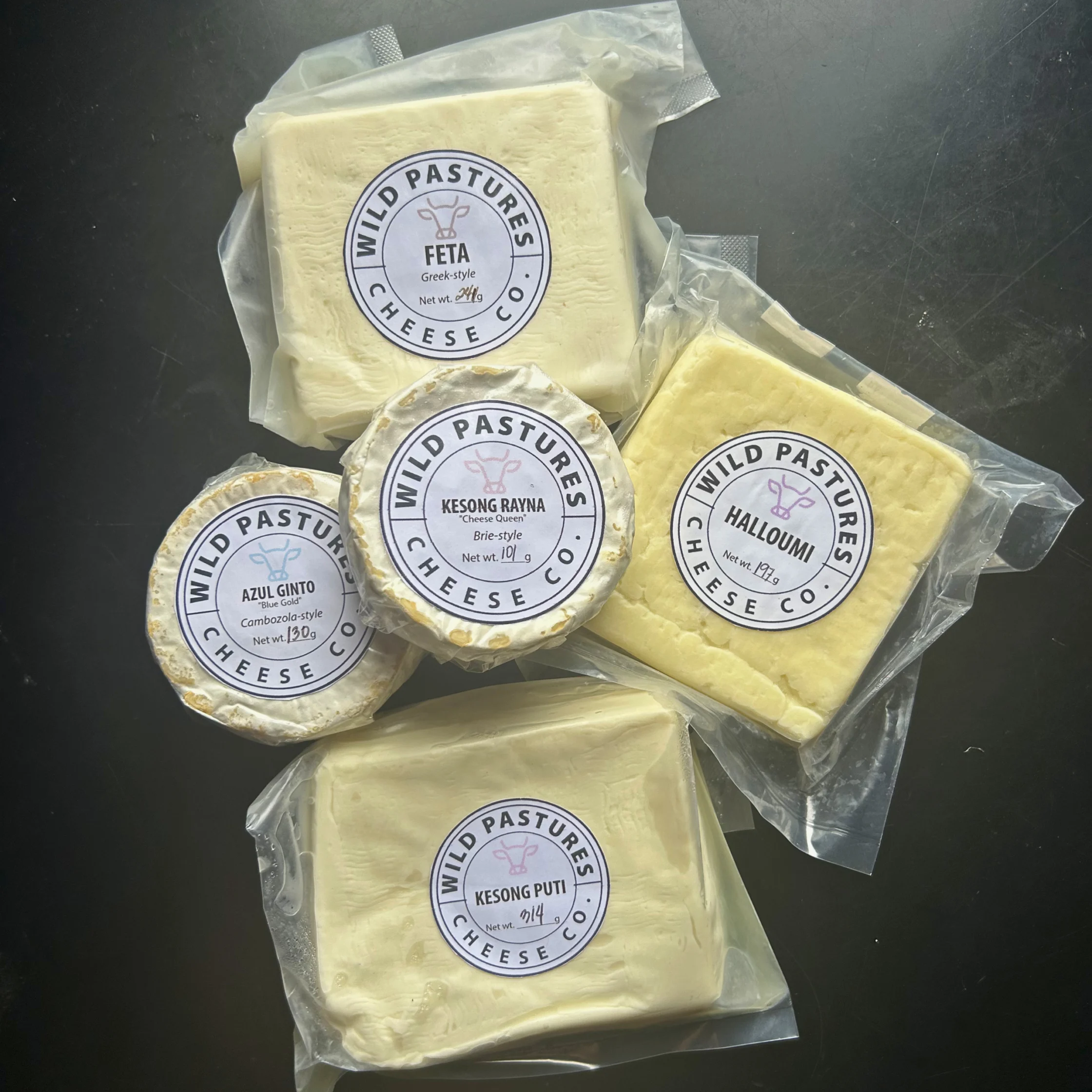 Assortment of Wild Pastures cheeses displayed in vacuum-sealed packaging, including feta, halloumi, brie-style, blue-style, and Kesong Puti cheeses from Wild Pasture Cheesemakers on a dark background.