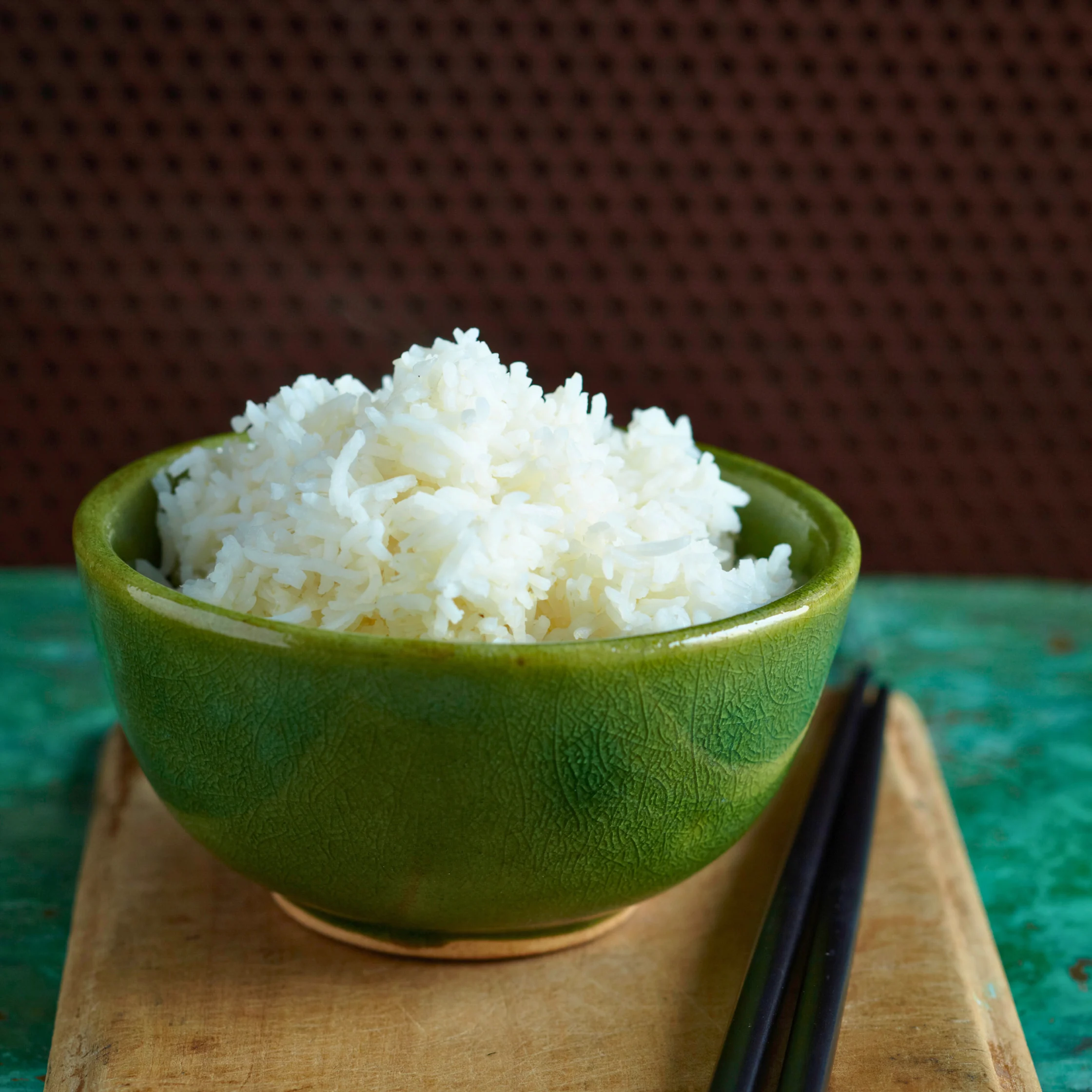 Green bowl filled with steamed white rice on a wooden board, with chopsticks beside it.
