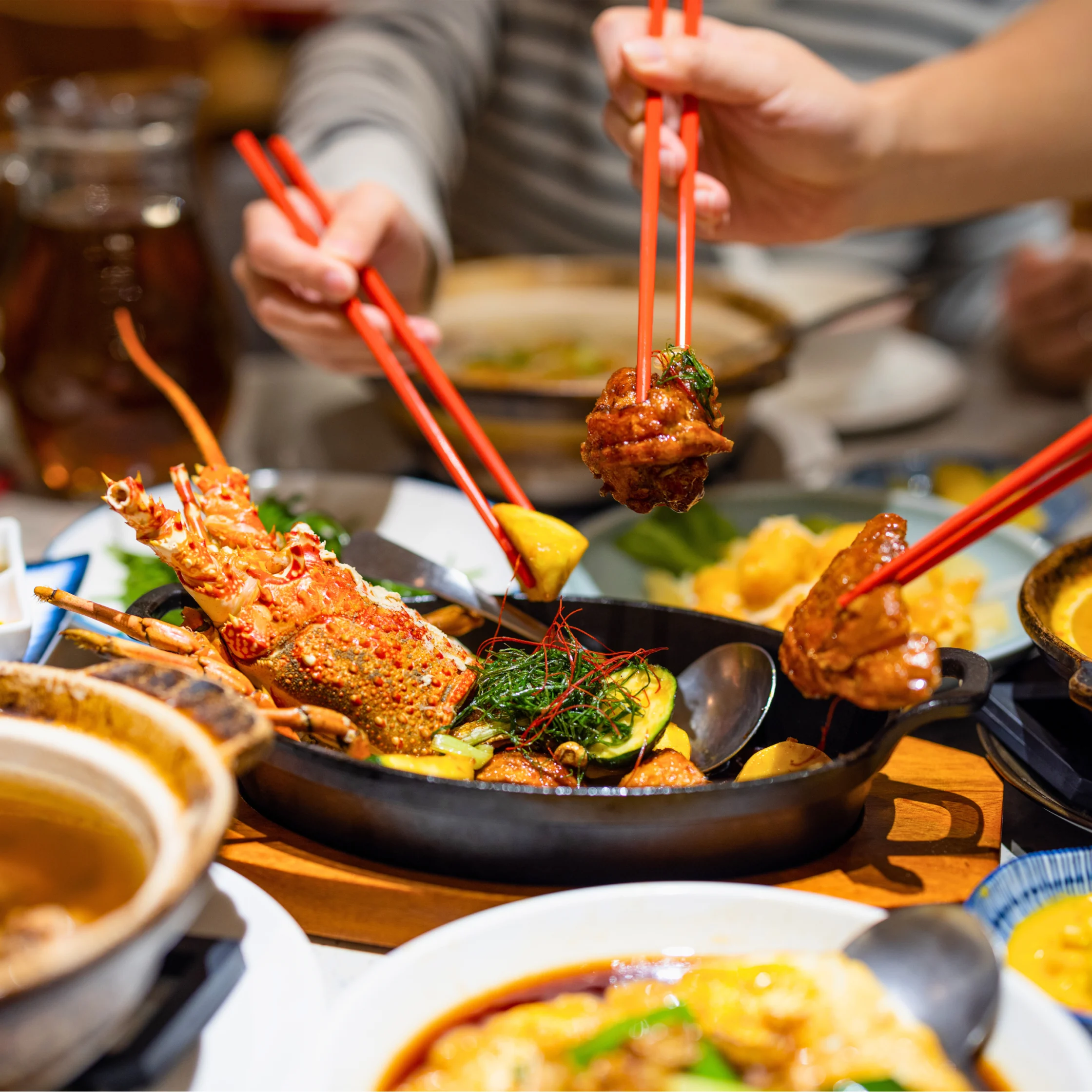 Diners using red chopsticks to share a seafood feast featuring lobster, vegetables, and grilled dishes served in cast-iron pans.