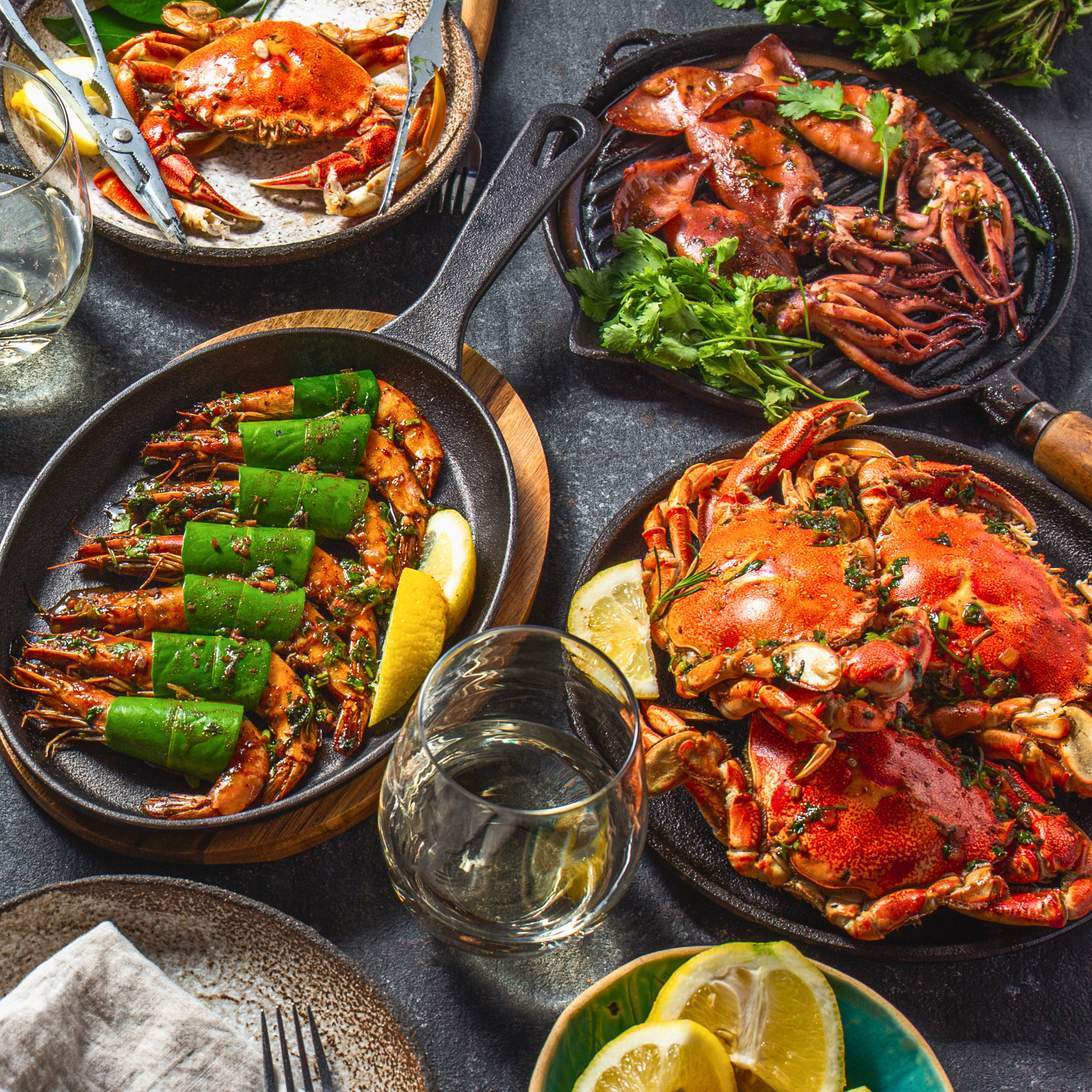 Overhead view of a seafood spread with crab, shrimp, squid, grilled prawns, lemon wedges, and wine served in cast-iron skillets.