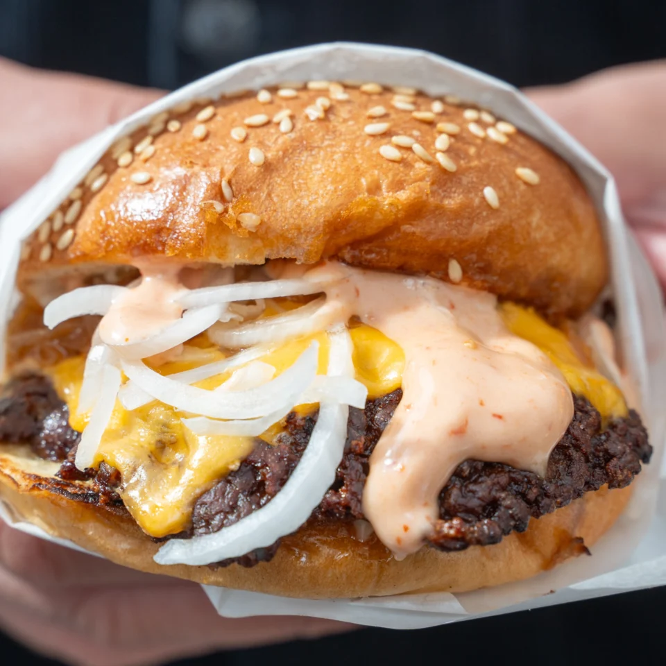 Close-up of a smash burger wrapped in paper with melted cheese, sliced onions, creamy sauce, and crispy-edged beef inside a toasted sesame bun.