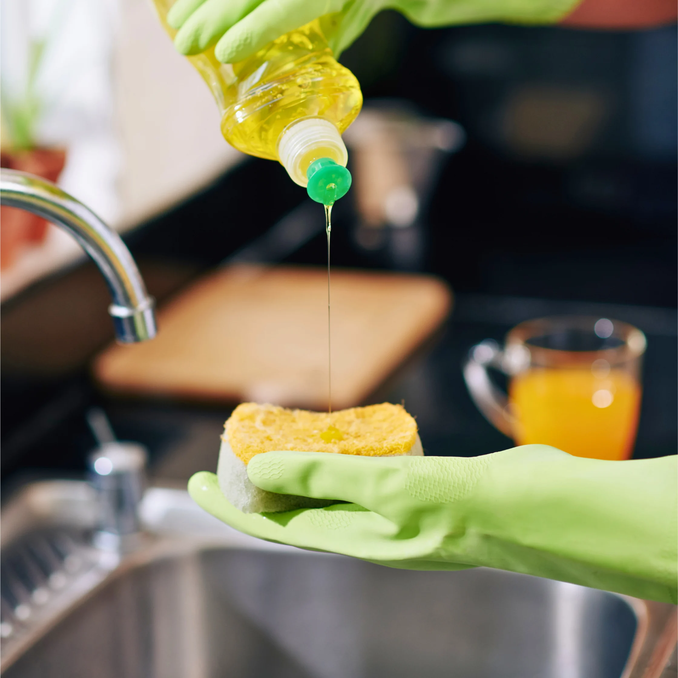 Person wearing cleaning gloves squeezing dish soap onto a kitchen sponge beside a sink.