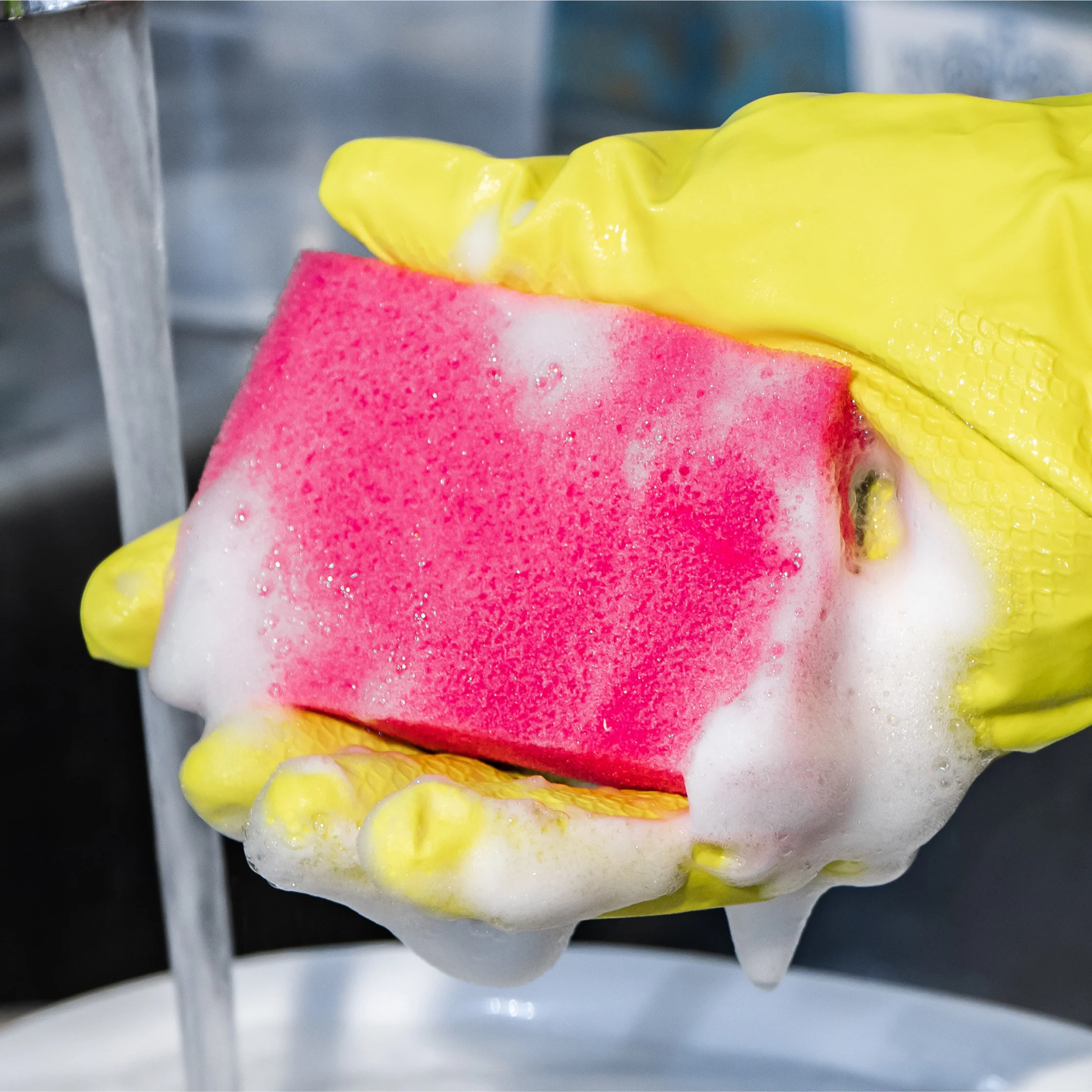 Gloved hands holding a pink sponge covered in soap suds beneath running water.