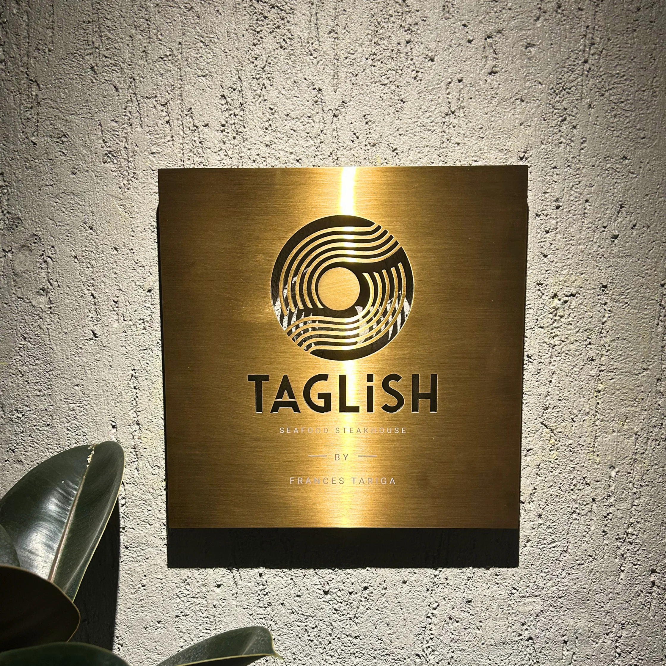 Gold wall plaque displaying the logo and name ‘Taglish Seafood Steakhouse by Frances Tariga.
