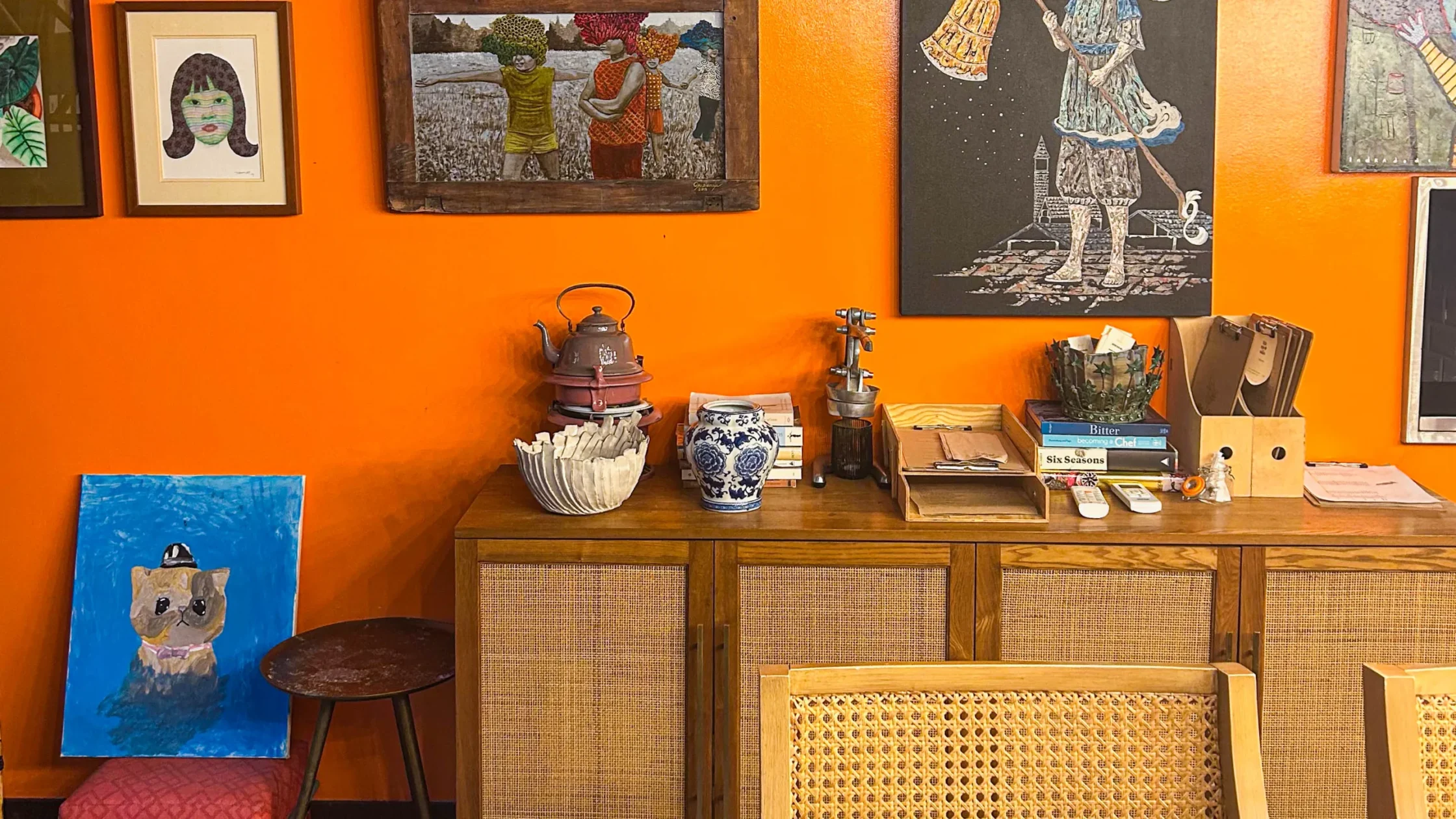 Warm dining room with a vivid orange wall displaying an eclectic collection of framed artworks above a wooden sideboard, complemented by woven chairs and patterned tile flooring.