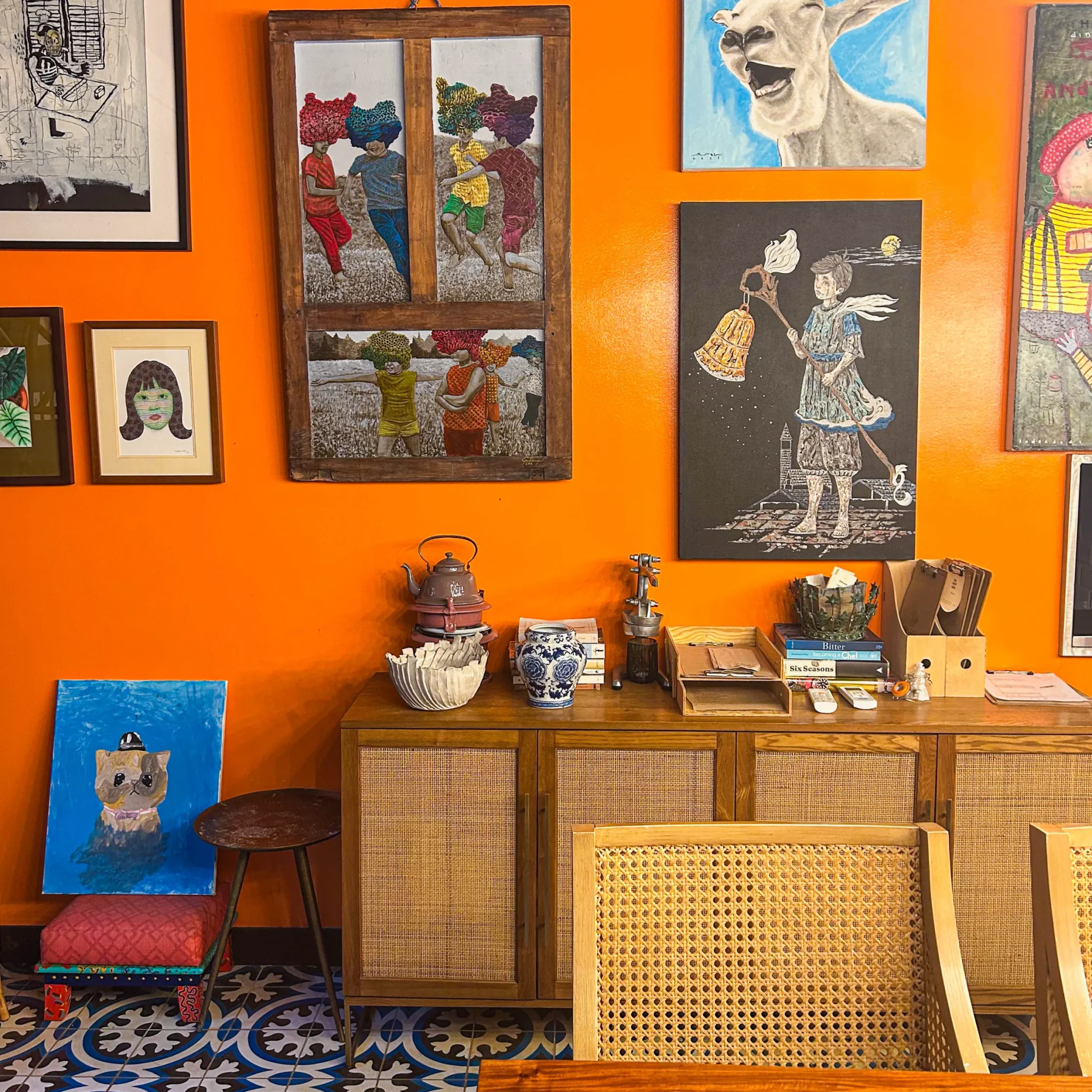 Warm dining room with a vivid orange wall displaying an eclectic collection of framed artworks above a wooden sideboard, complemented by woven chairs and patterned tile flooring.