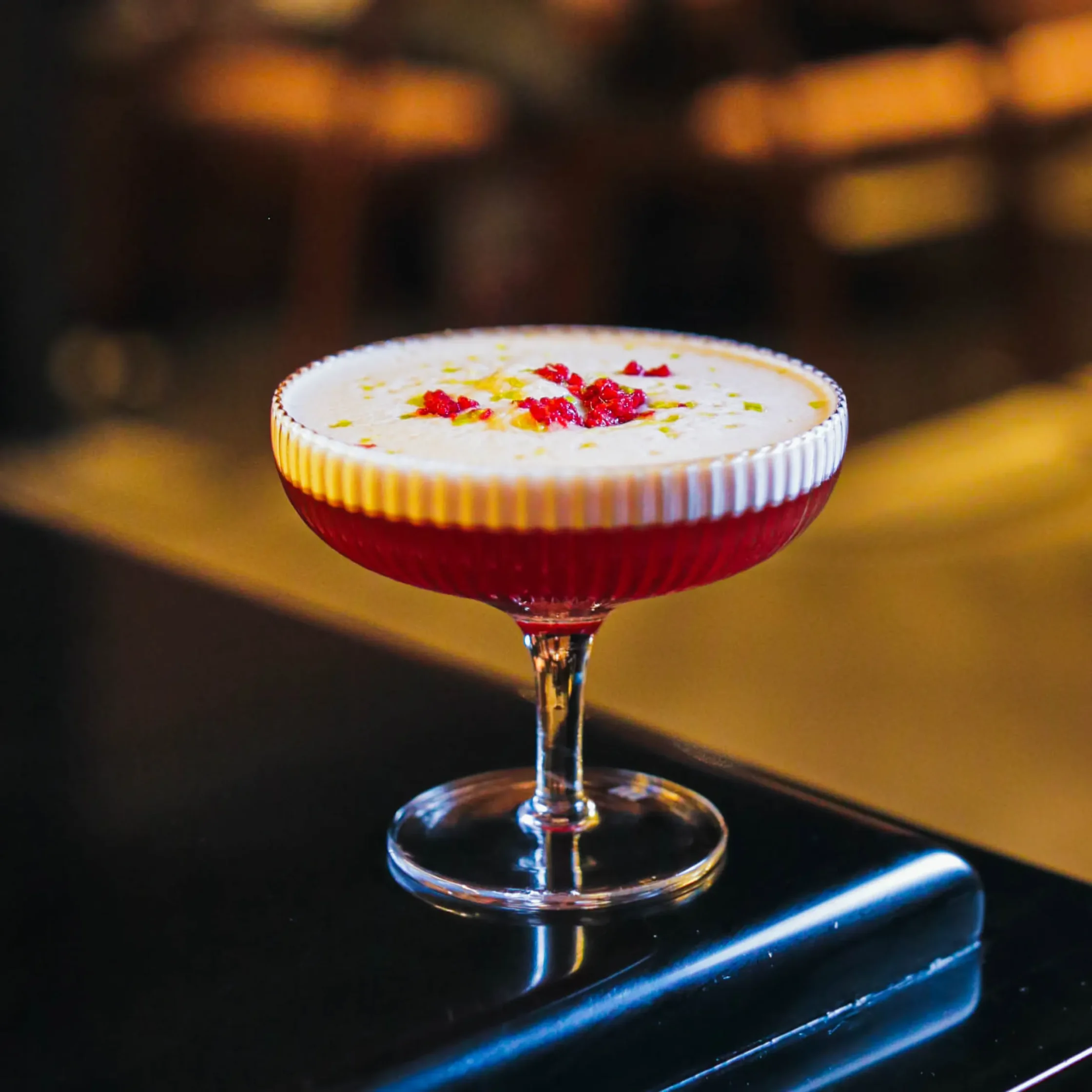 A bright red cocktail topped with a thick white foam and red garnish, served in a coupe glass on a bar counter at Toma Toma in Makati.