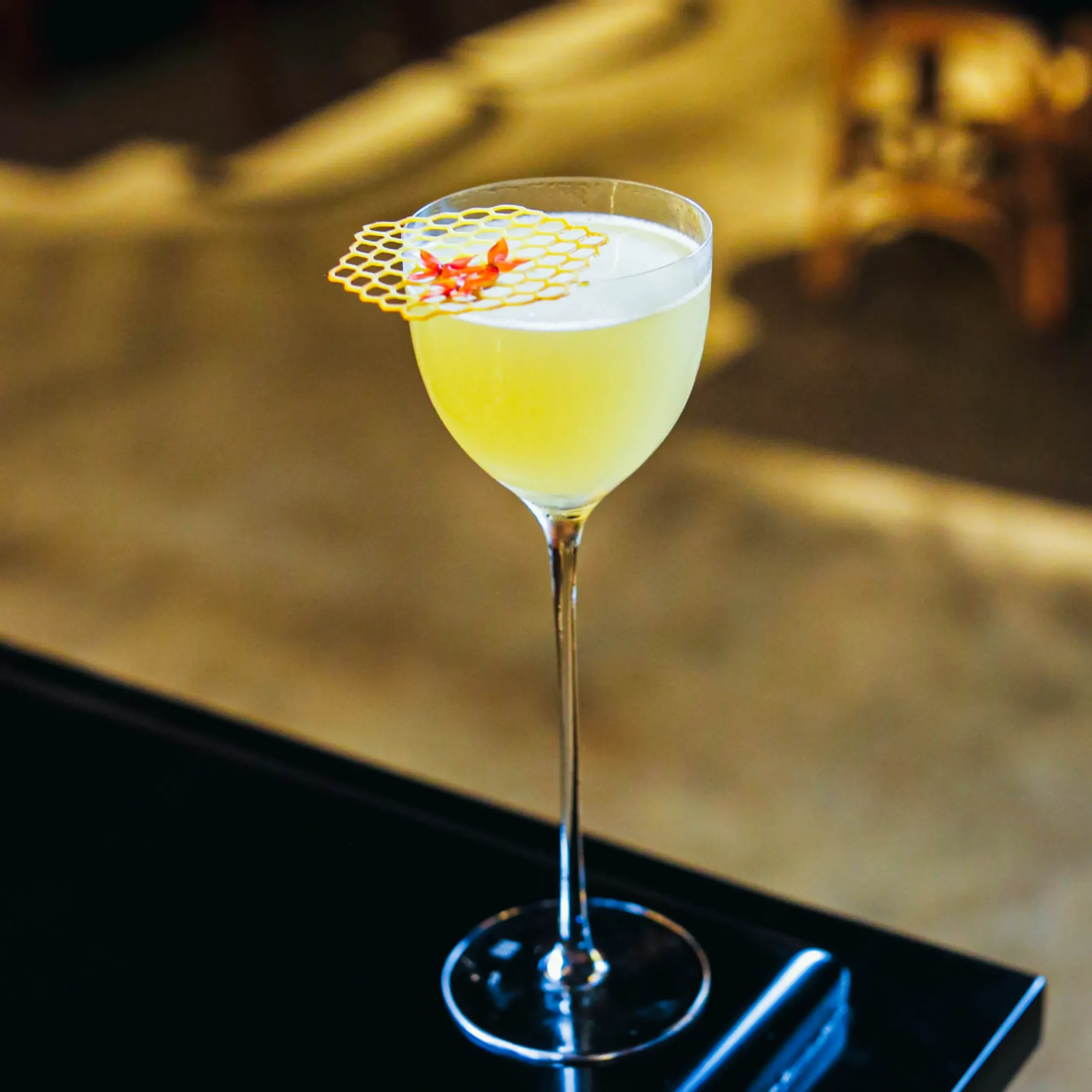 A pale green cocktail in a tall stemmed glass, garnished with a honeycomb-pattern crisp and edible flowers, highlighting Toma Toma's use of local Philippine ingredients.