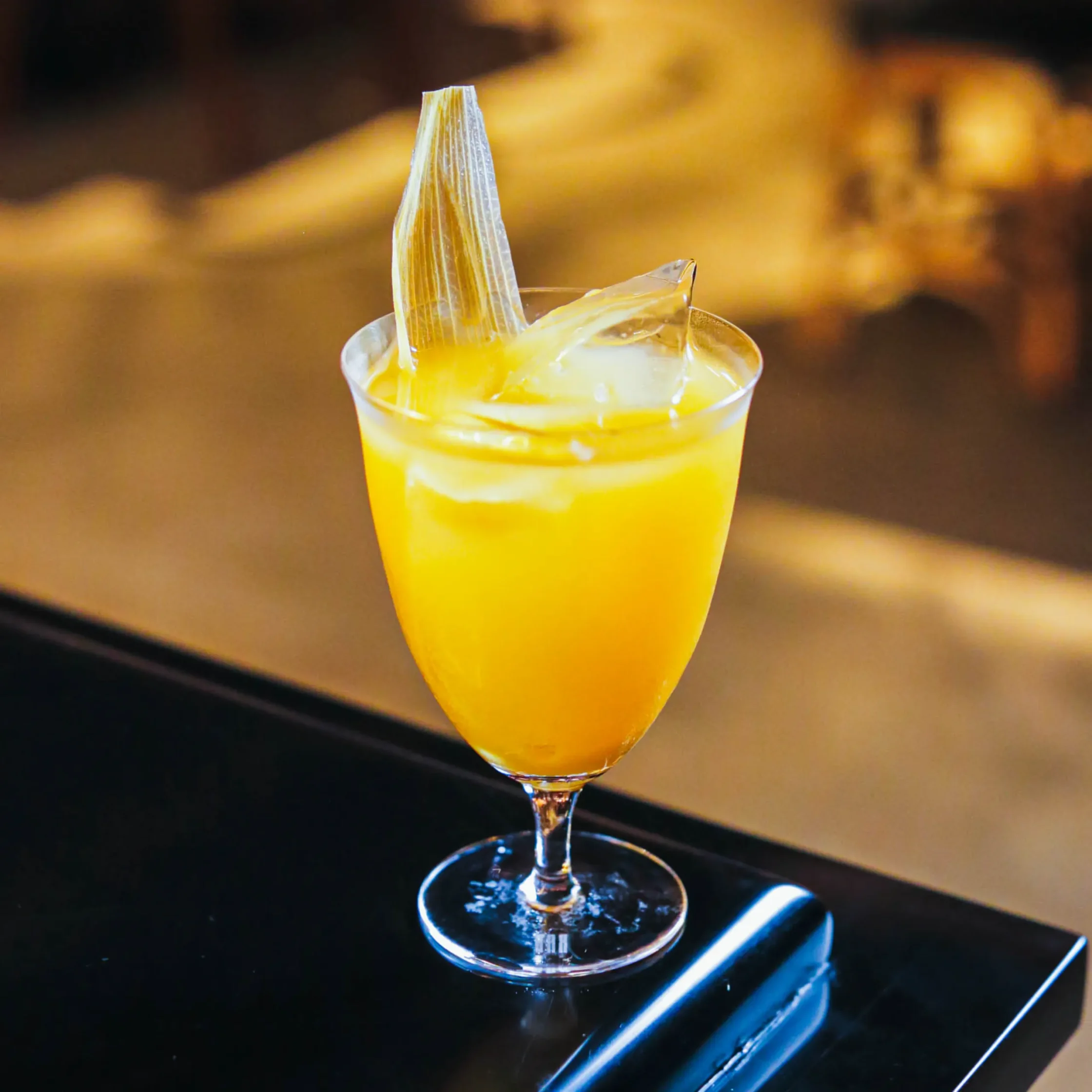 A vibrant yellow cocktail served over large ice cubes in a stemmed glass, garnished with a delicate translucent crisp at Toma Toma cocktail salon.