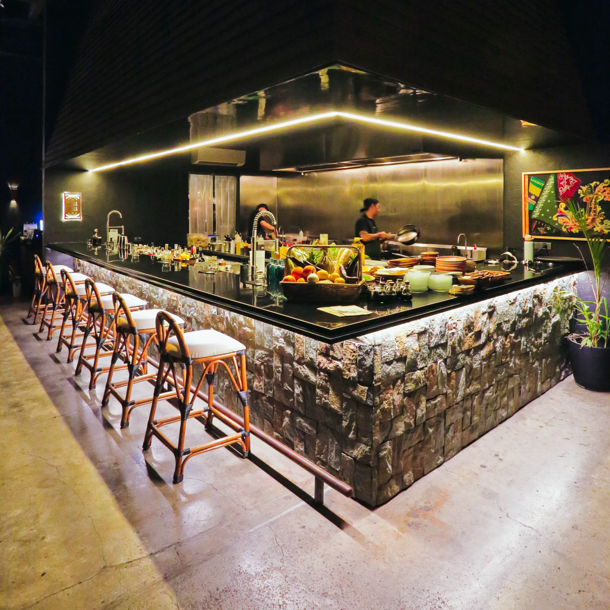 The open bar at Toma Toma features stone-clad counters, high stools, warm lighting, and a visible kitchen where guests can watch food and drinks being prepared.