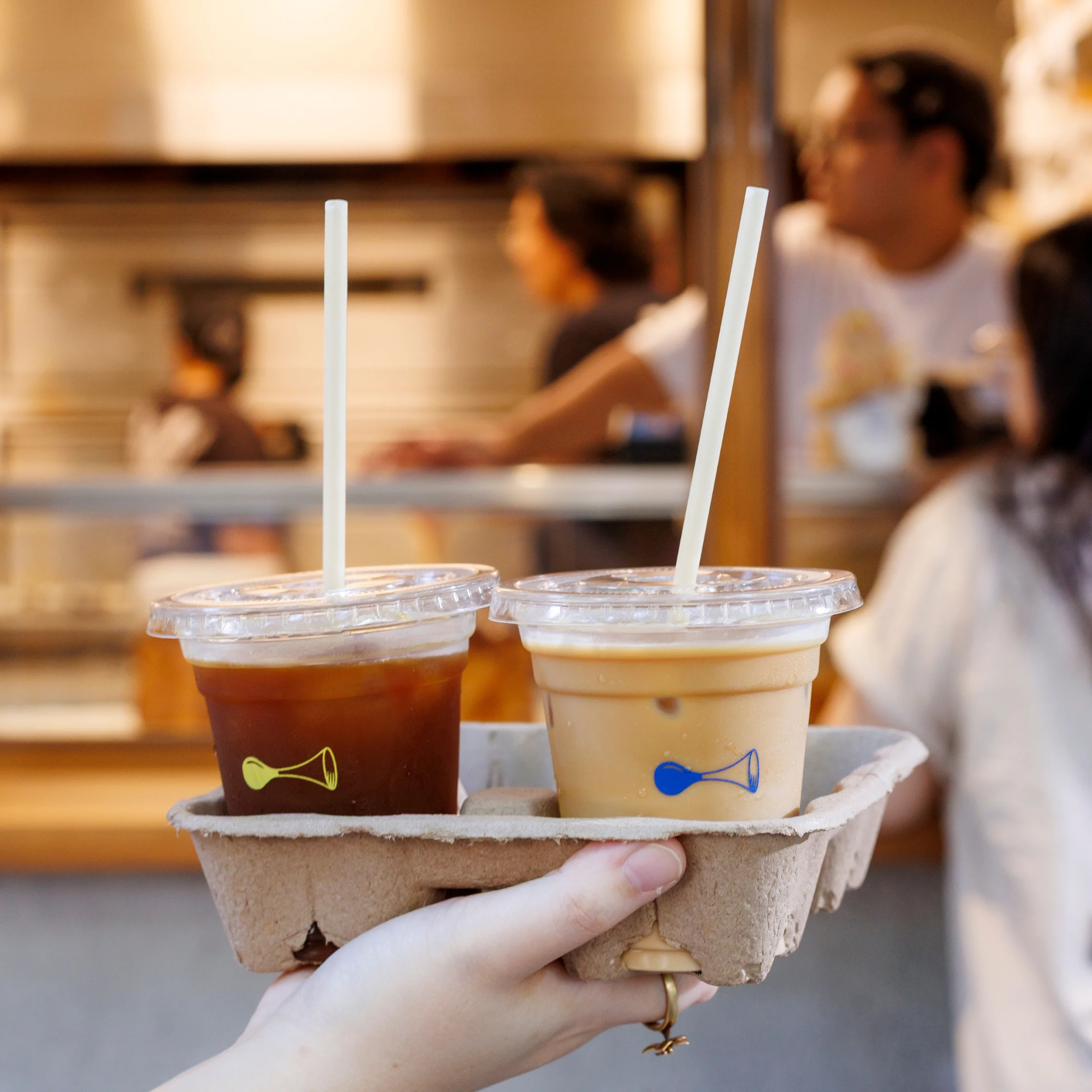 Two iced drinks in clear cups—one dark and one creamy—held in a cardboard carrier in front of a softly blurred café setting with people in the background.
