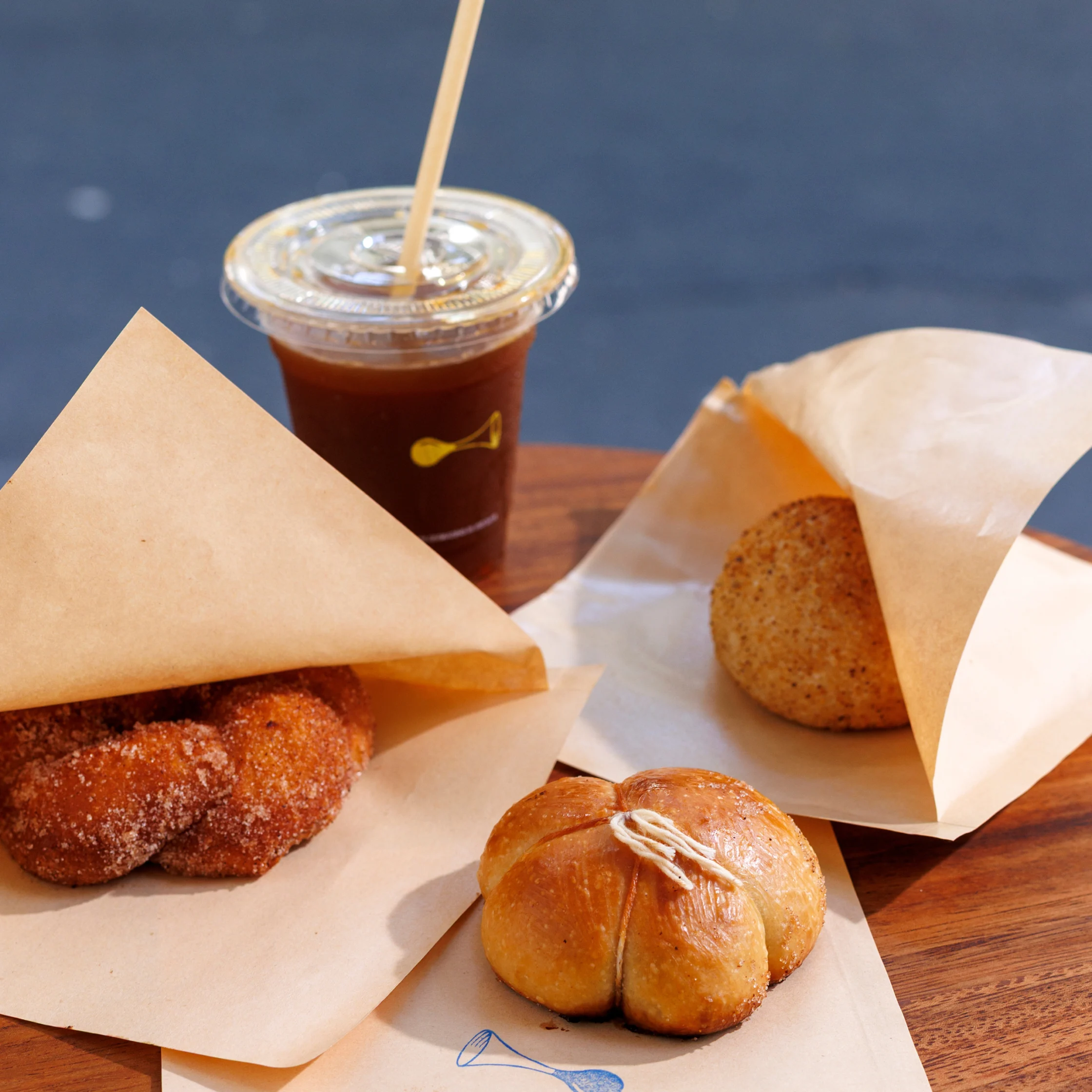 An assortment of pastries and breads arranged on a wooden table beside an iced drink, with baked items partially wrapped in paper for serving.