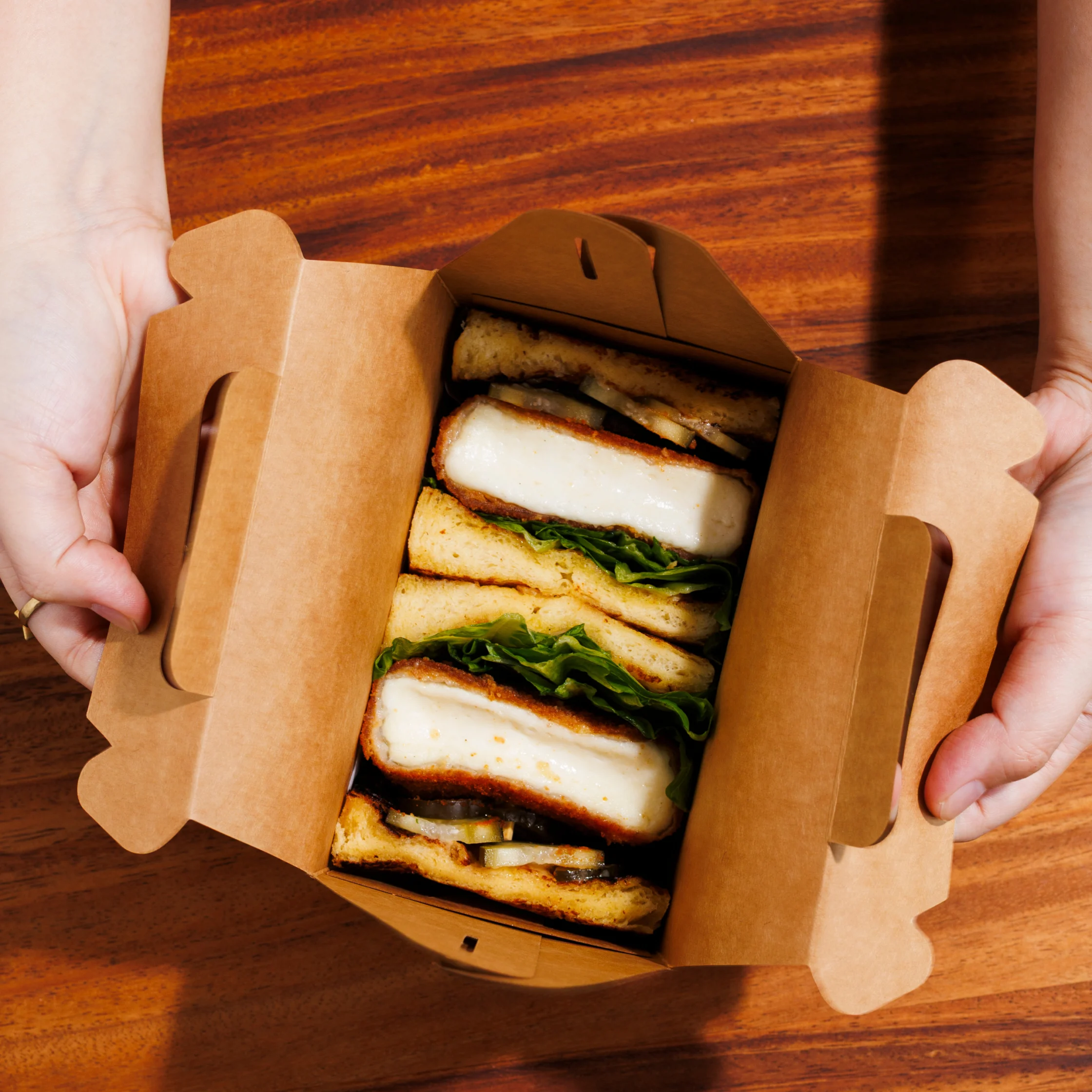 Open cardboard takeout box filled with stacked sandwiches featuring thick breaded fillings, leafy greens, and sliced vegetables, held above a wooden tabletop.