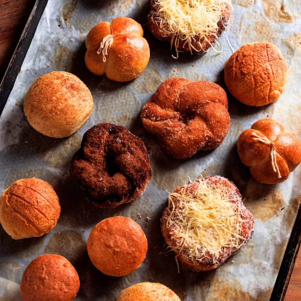 Overhead spread of assorted pastries and breads on a parchment-lined tray, including sugar-coated, chocolate-dusted, and cheese-topped varieties.