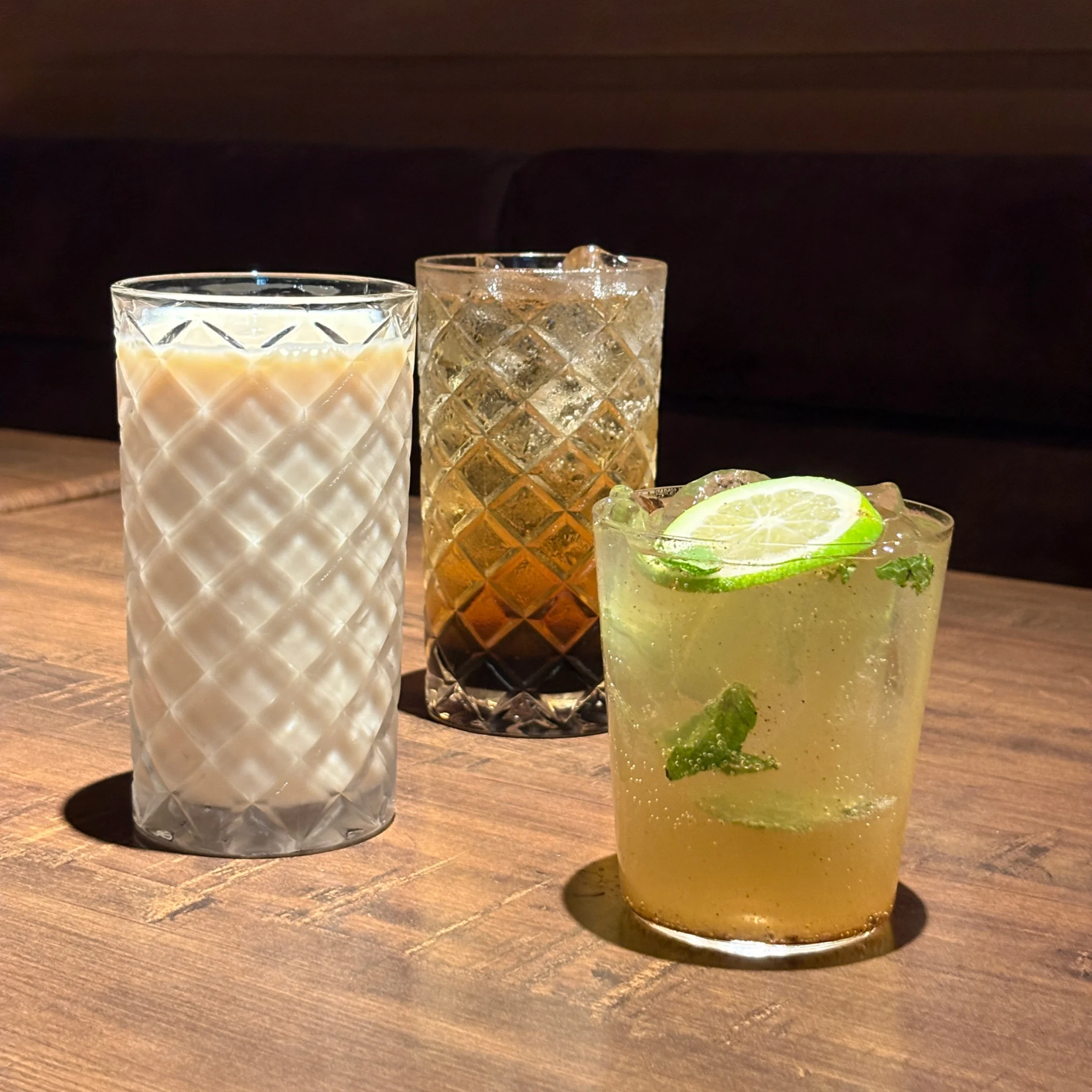 Three drinks on a wooden restaurant table: a creamy cold soy milk in a tall glass, a dark iced gulaman drink in a patterned glass, and a sparkling virgin mule garnished with lime and mint.