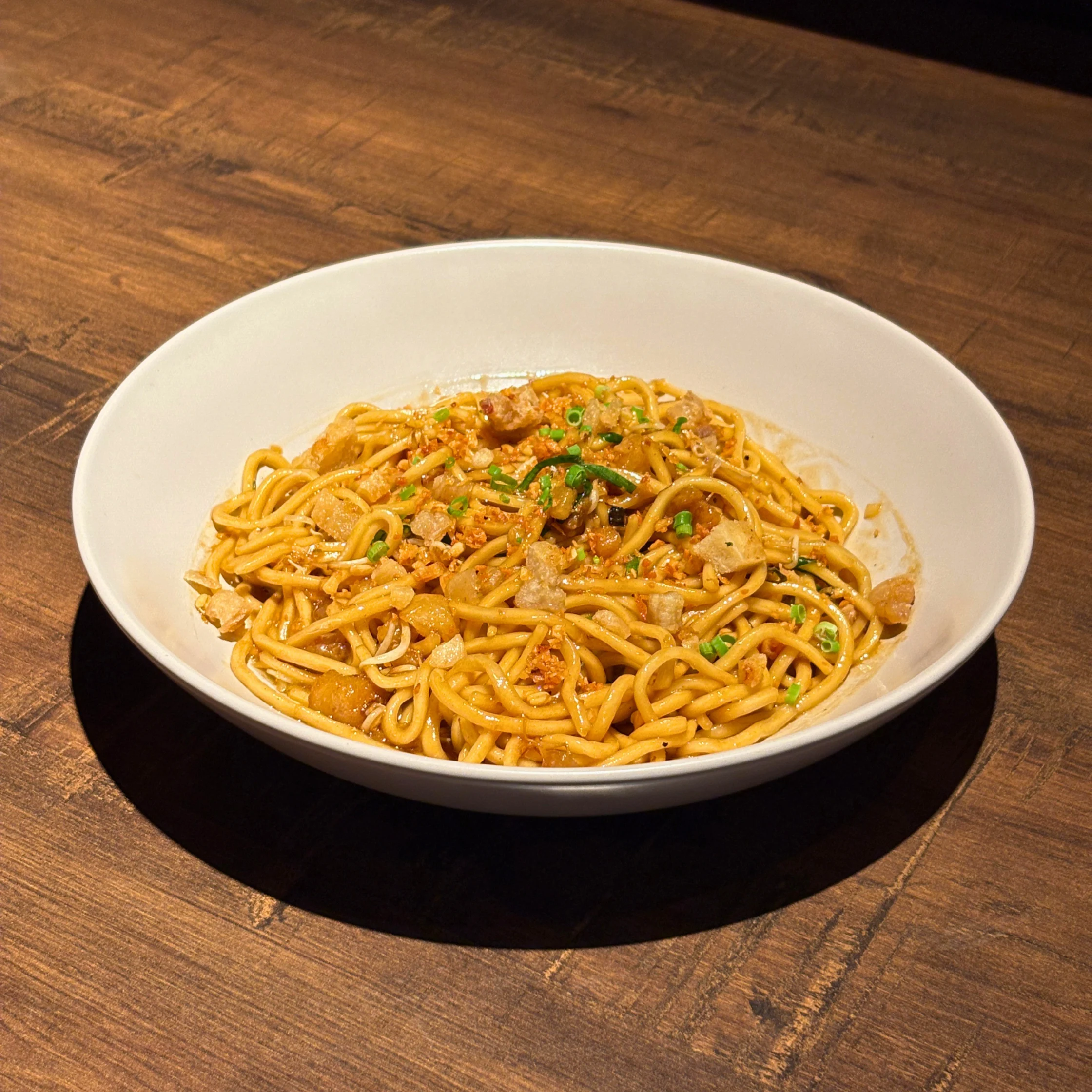 Bowl of garlic butter noodles topped with crispy garlic pieces and chopped green onions, served on a wooden table.