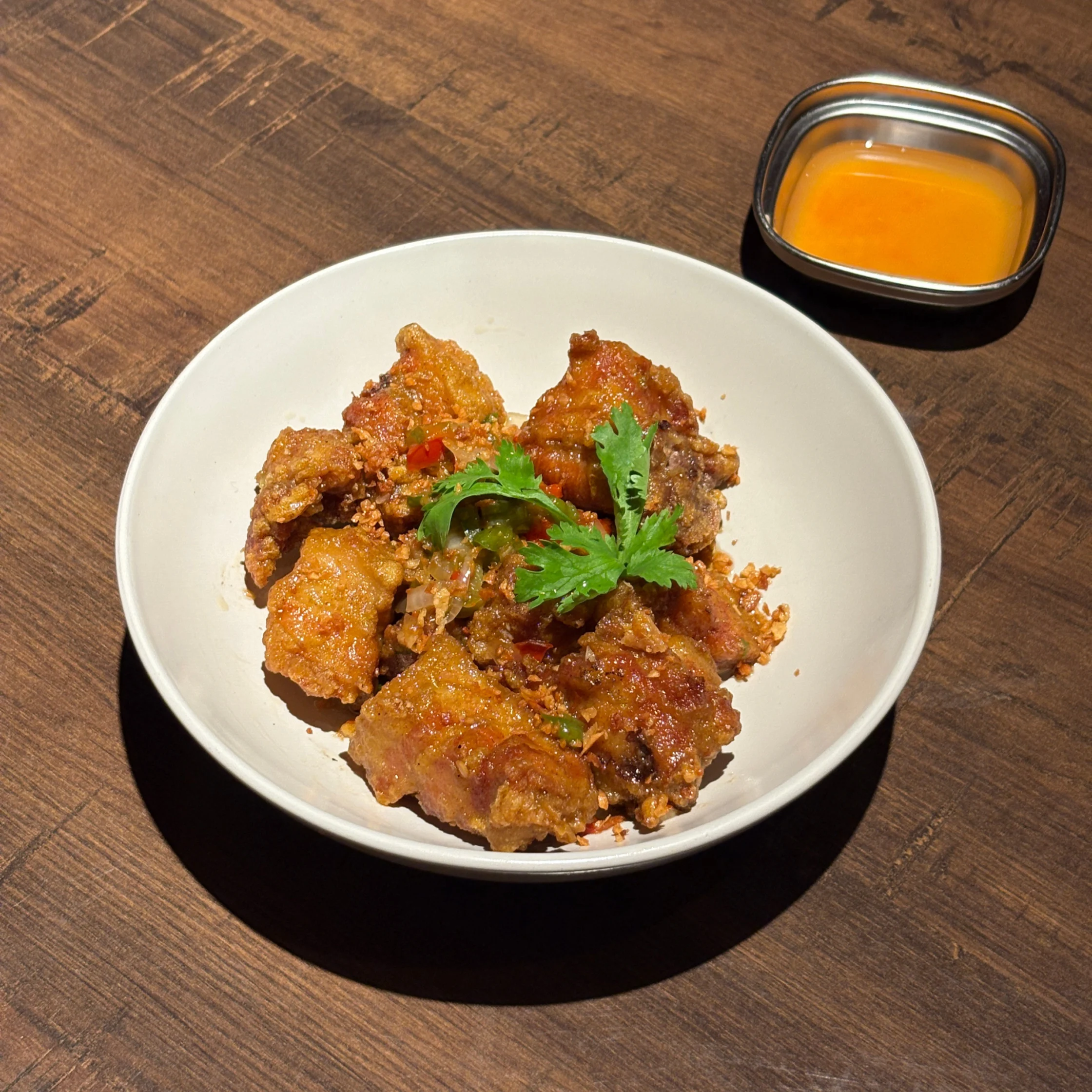 Bowl of crispy salt-and-pepper spare ribs garnished with cilantro, sliced peppers, and fried garlic, with a side of dipping sauce.