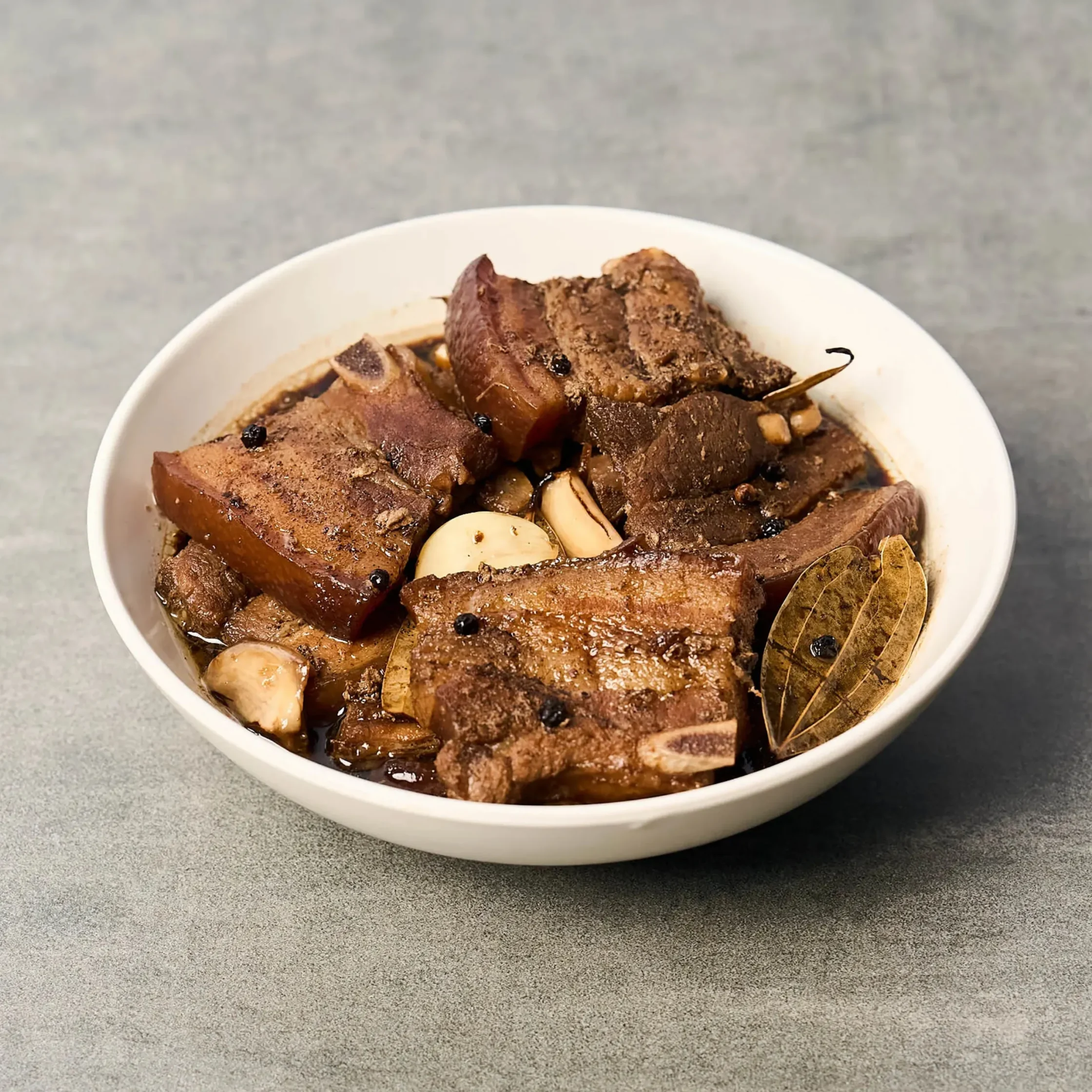 Close-up of a white bowl filled with braised beef slices, studded with peppercorns, with garlic and a bay leaf on the side.