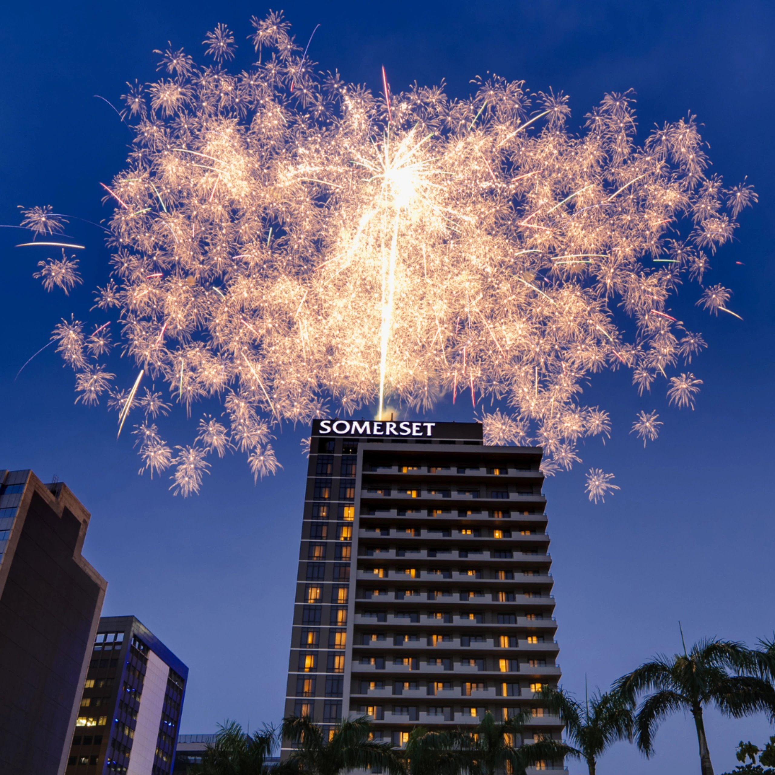 Tall modern hotel building with a bright 'SOMERSET' sign, fireworks lighting the night sky above.