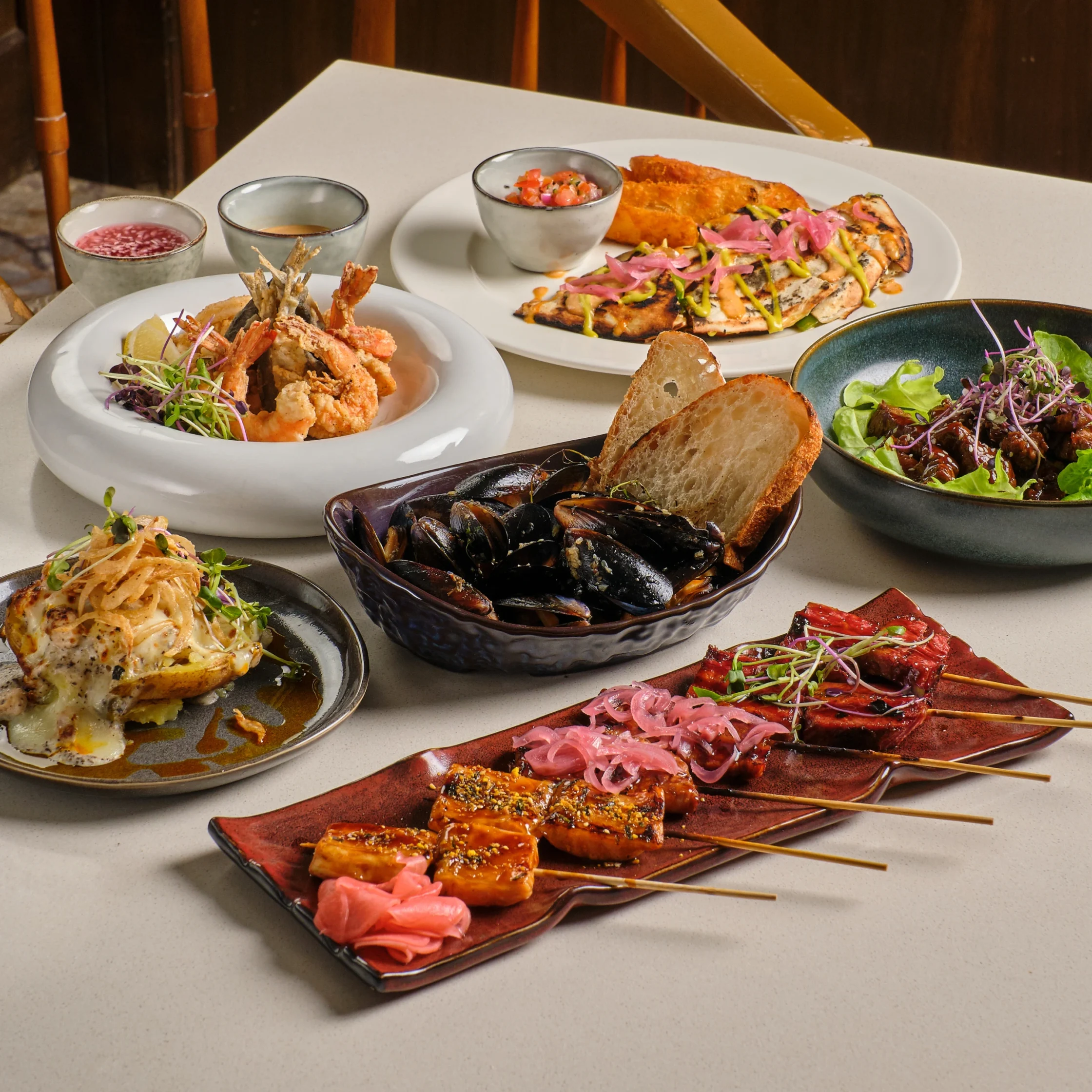 Assorted plated dishes on a table: fried shrimp tempura, skewered grilled meat with pink onions, mussels, bread, and small sauce bowls.