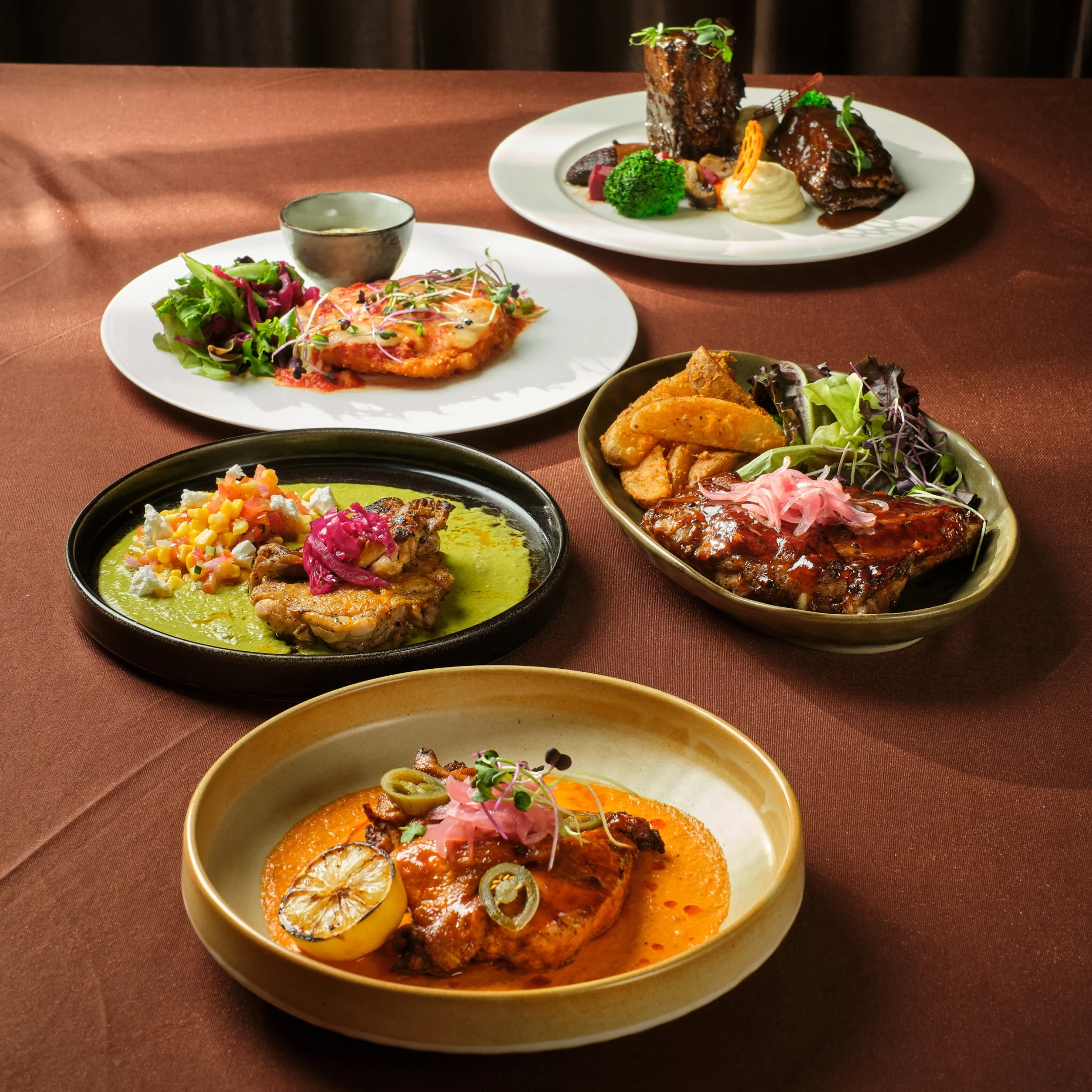 Assorted plated dishes on a brown table: a colorful salad, a bowl with green sauce and toppings, and a plate with braised meat in dark glaze.