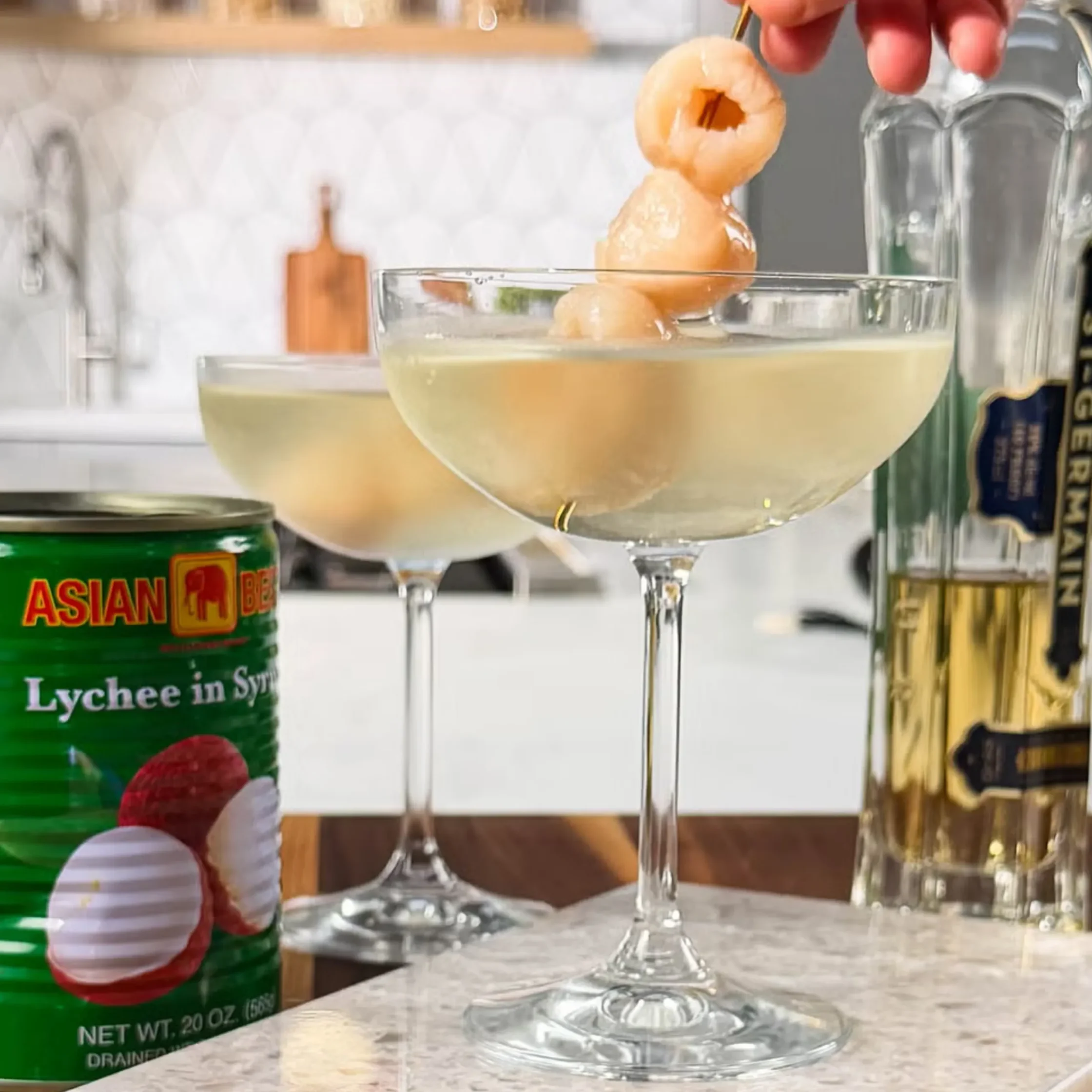 Cocktail glass with a lychee skewer being dipped into a pale drink, a green can of lychee drink visible in the foreground.