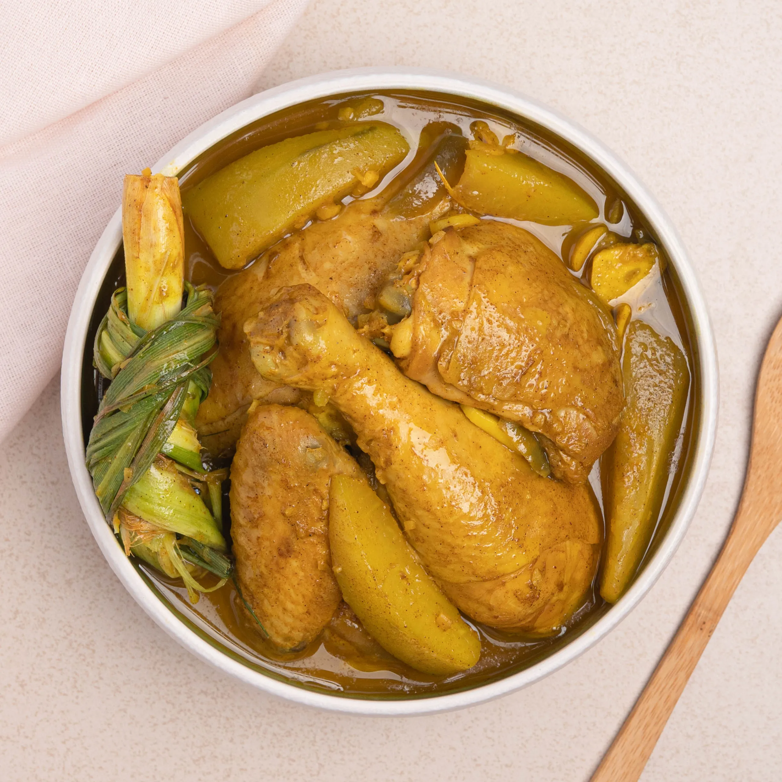 Top-down view of braised chicken with plantains in a thick yellow-brown sauce, with a wrapped leafy bundle beside it in a white dish.